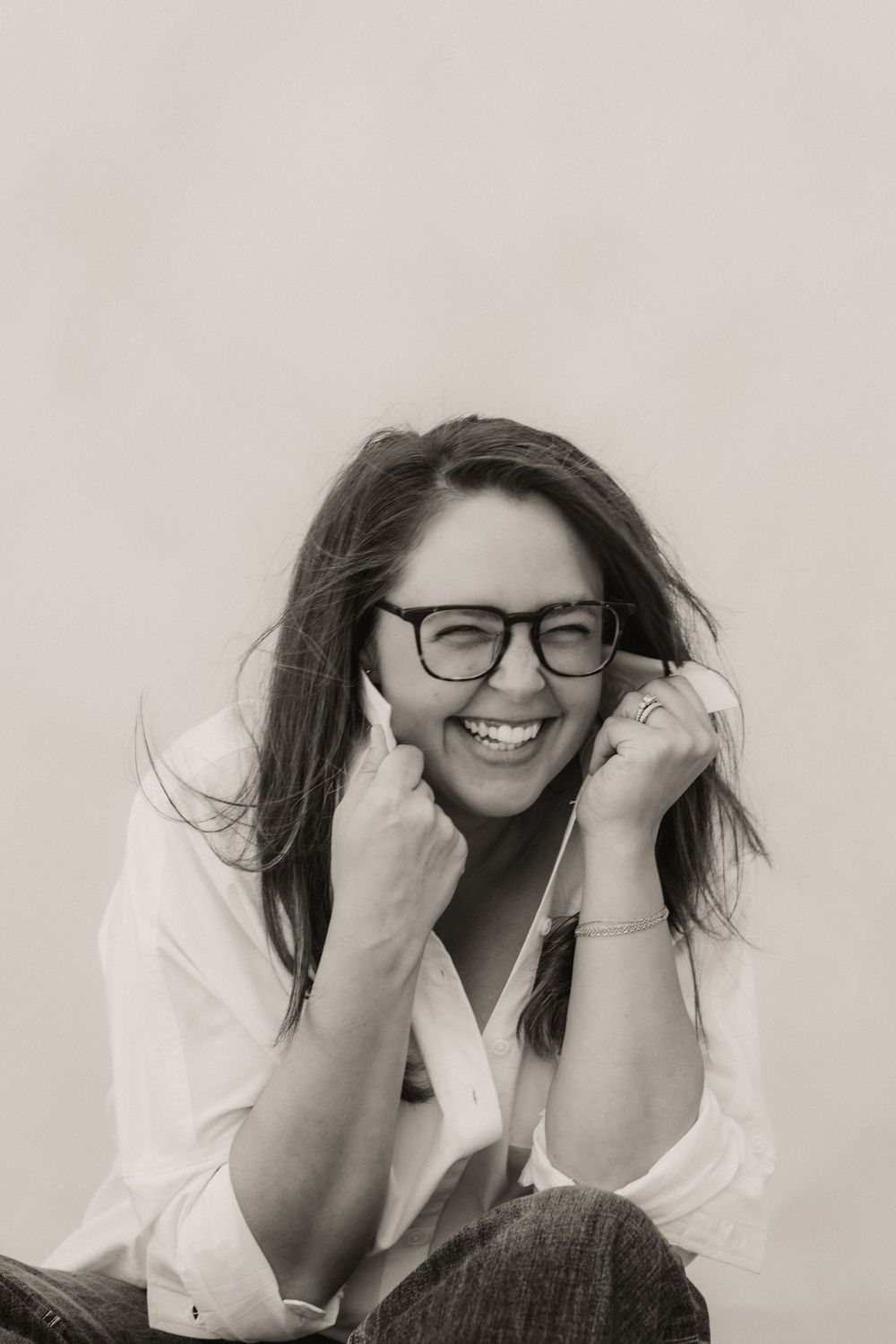 Black and white photo of a laughing woman with glasses and long hair, wearing a white shirt, posing casually.