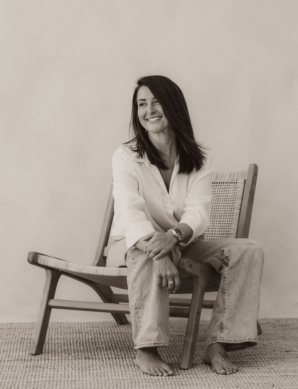 A smiling woman with dark hair sits barefoot in a wooden cane chair, wearing a white blouse and jeans.