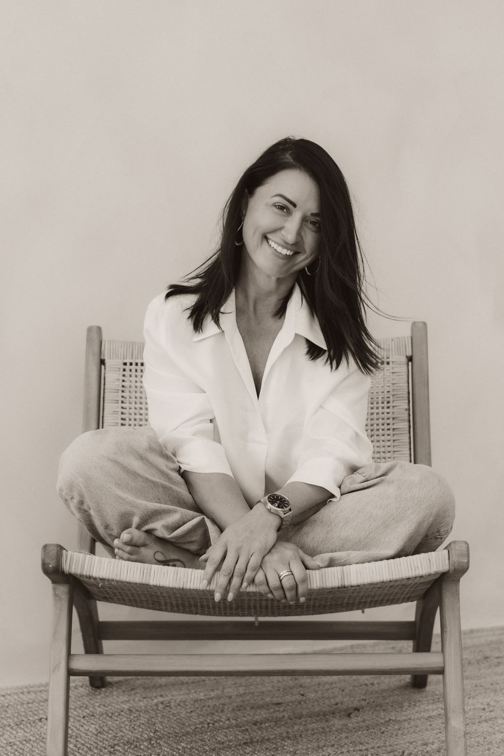 A smiling woman with dark hair sits cross-legged on a wooden chair, wearing a white shirt and jeans, in a sepia-toned photo.