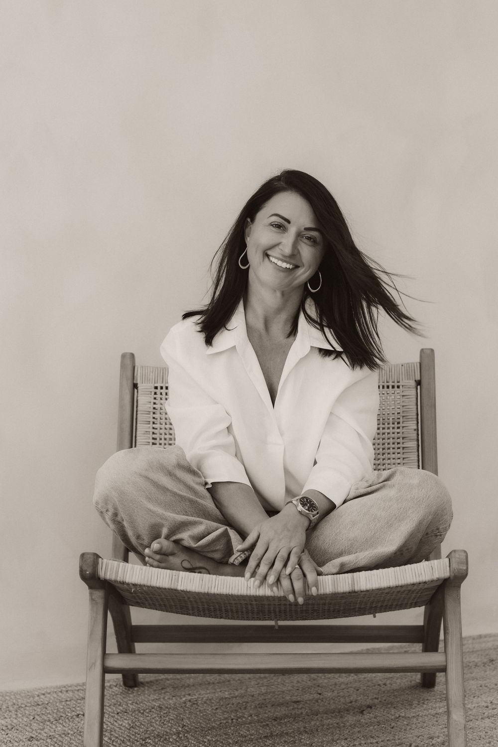 Smiling woman with dark hair sits cross-legged on a wooden chair, wearing a white shirt and jeans, in a black and white photo.