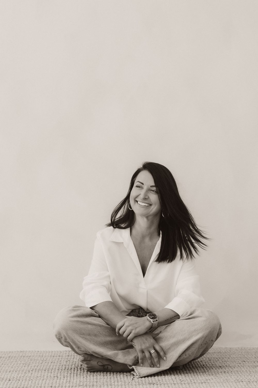 Black and white photo of a smiling woman with dark hair sitting cross-legged on the floor in a white shirt.