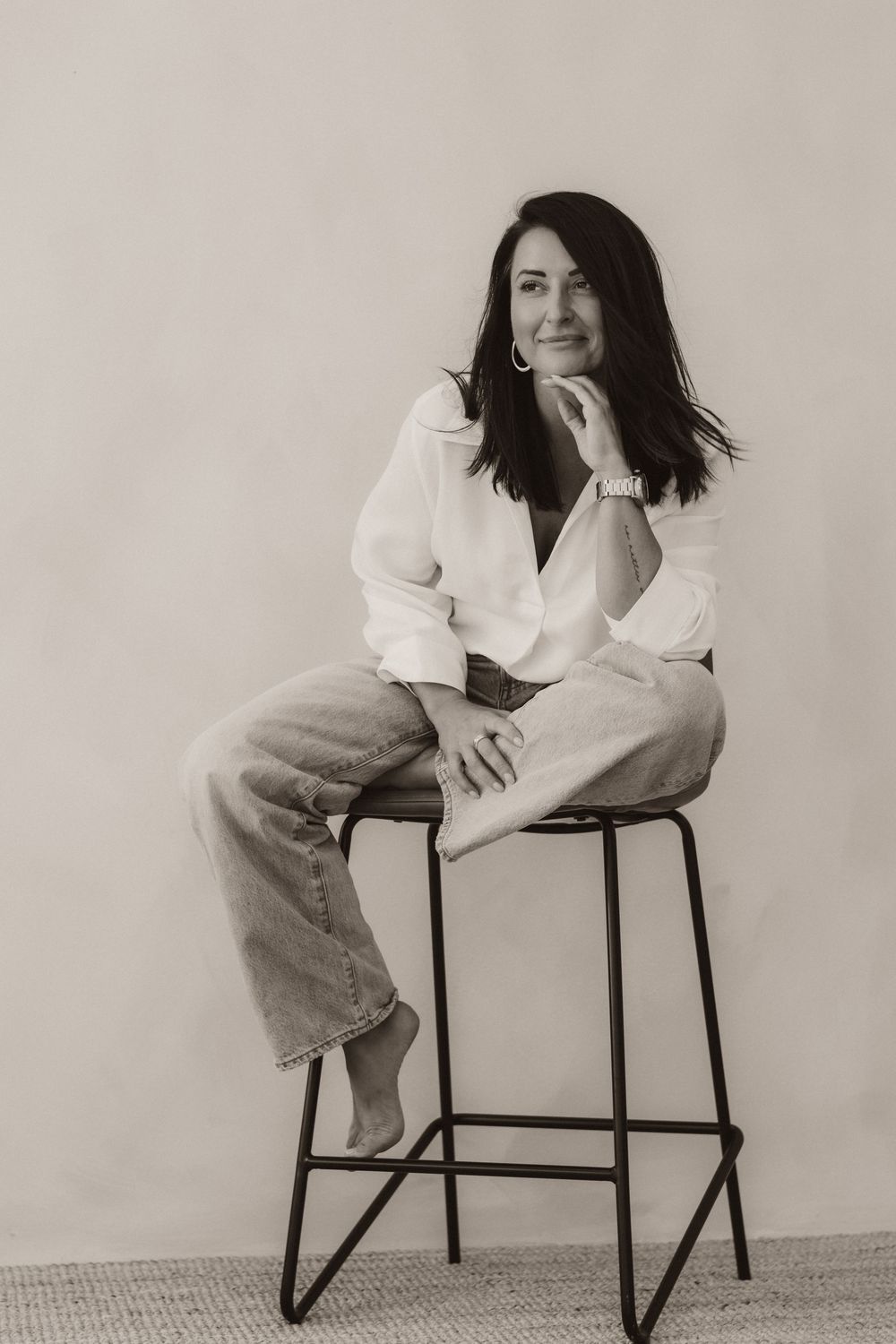 Black and white photo of a woman sitting on a stool in white shirt and jeans, posing with hand on chin.