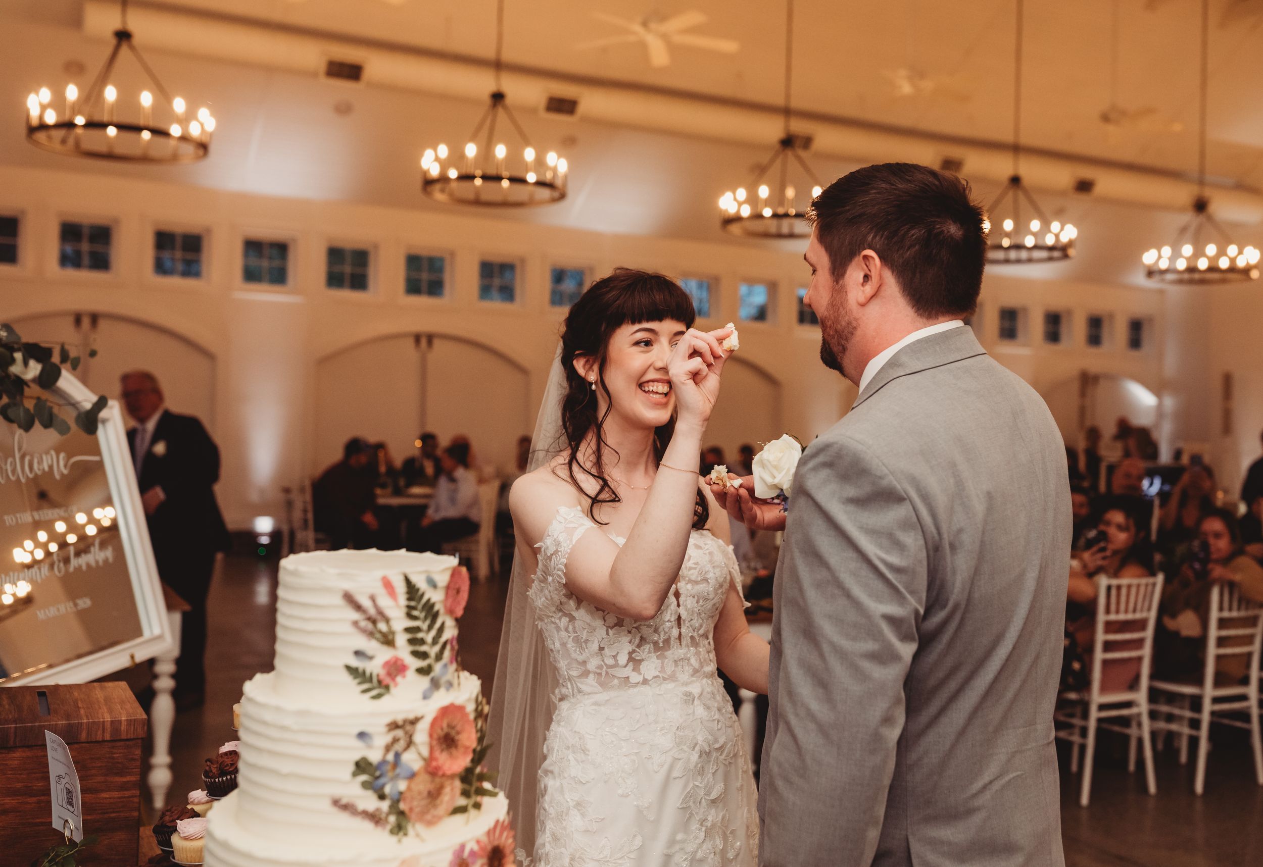 Bride feeding groom wedding cake during reception in elegant ballroom with chandeliers and white tiered cake.