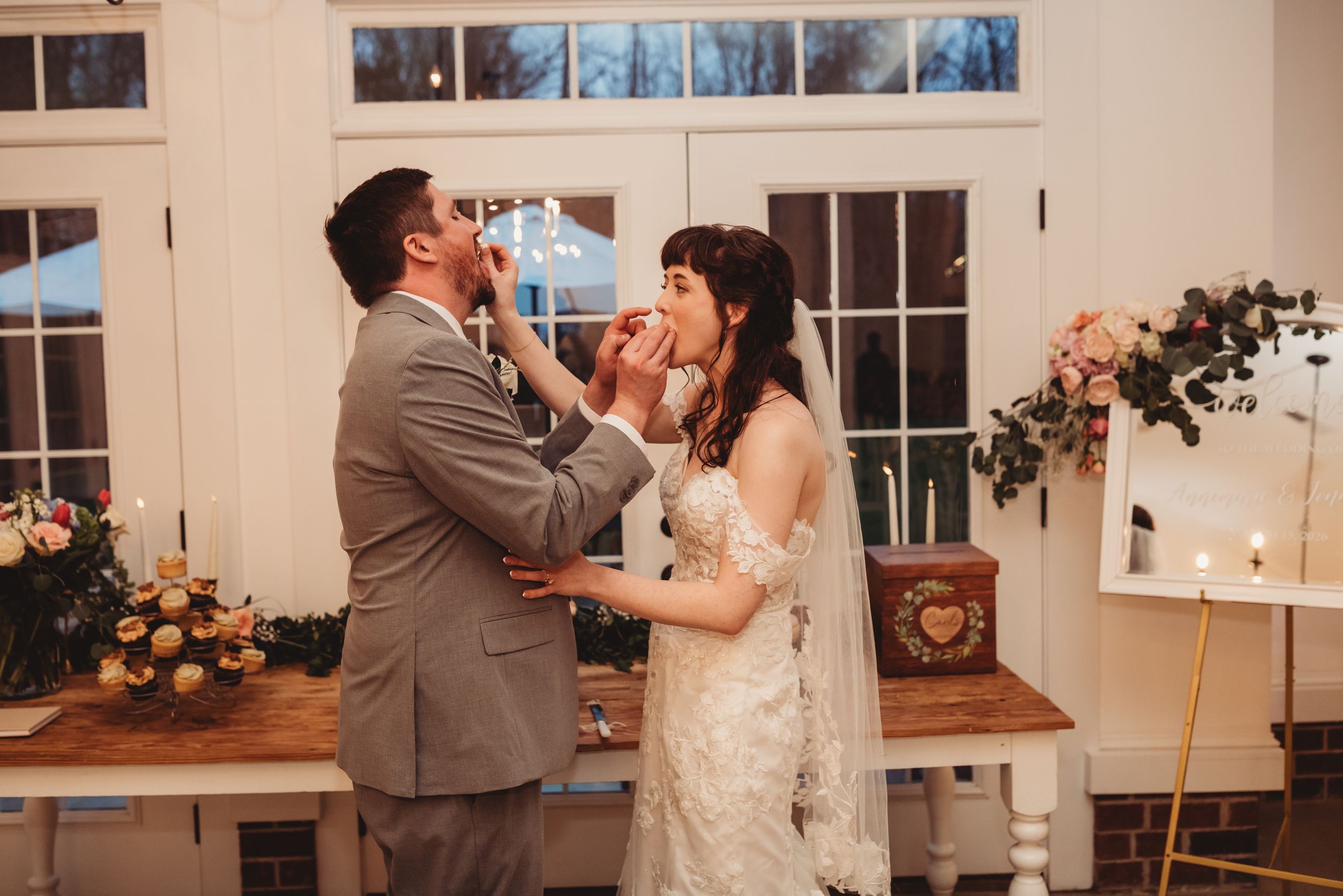 Bride and groom feeding each other wedding cake at elegant indoor reception with floral decor and candles.
