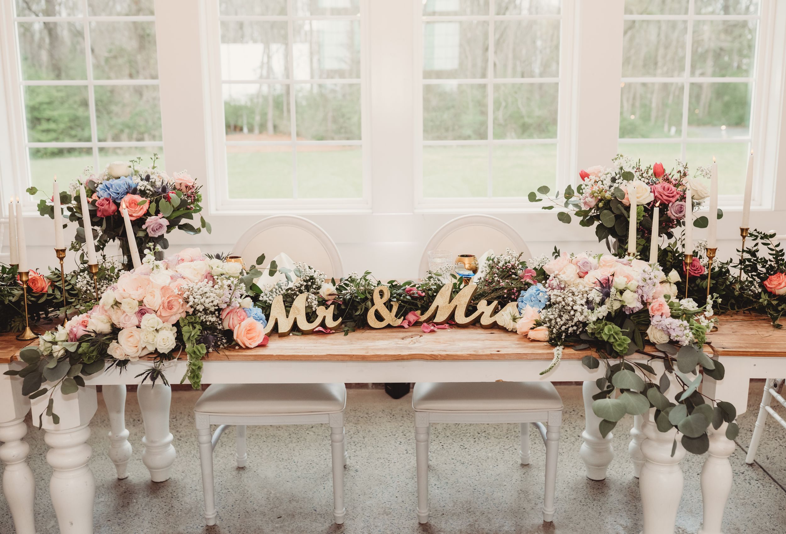 Elegant wedding sweetheart table with Mr & Mrs sign, lush floral garland of pink and white roses, and candles.