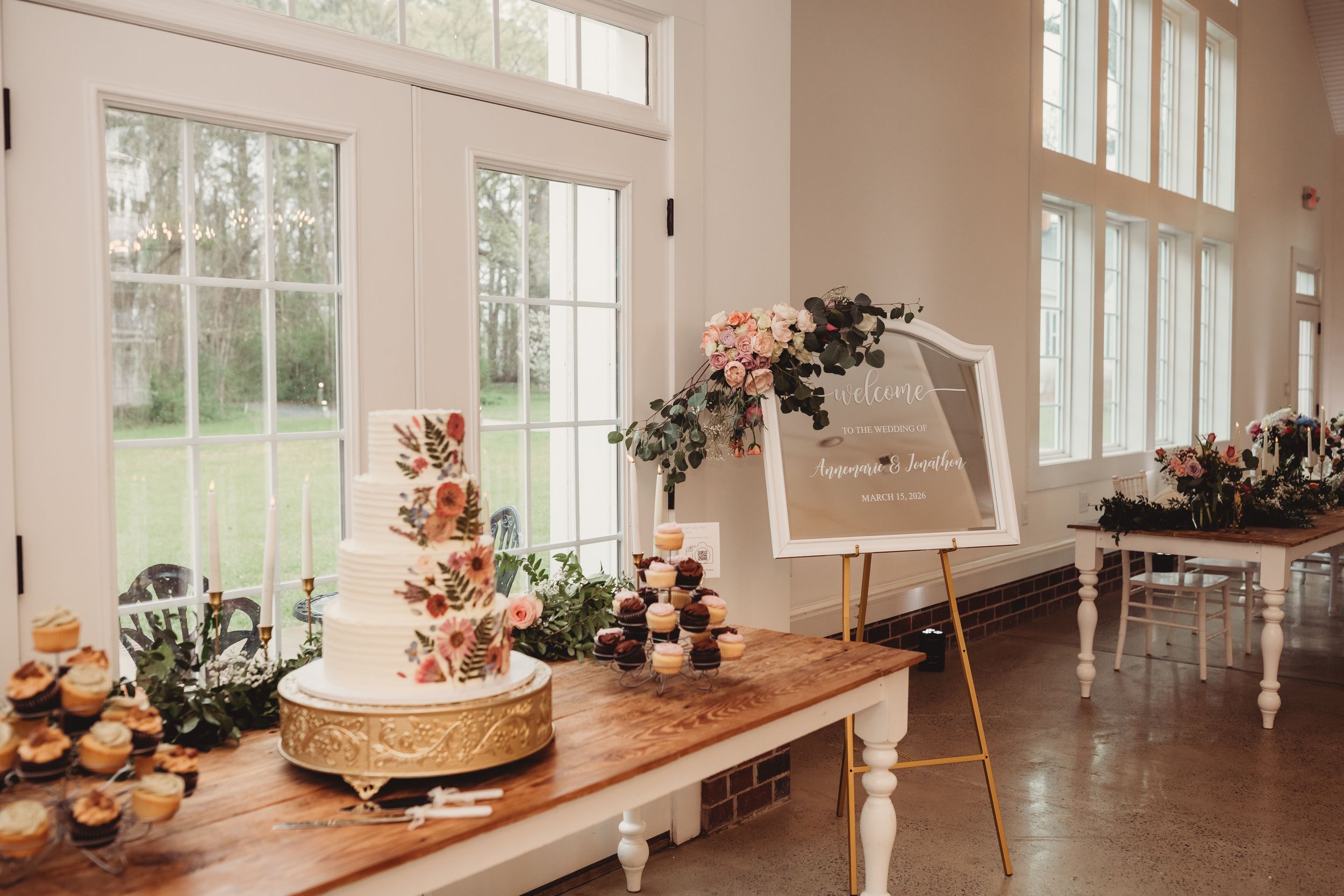 Elegant wedding dessert table with tiered cake on gold stand, cupcakes, and floral welcome sign in bright venue.