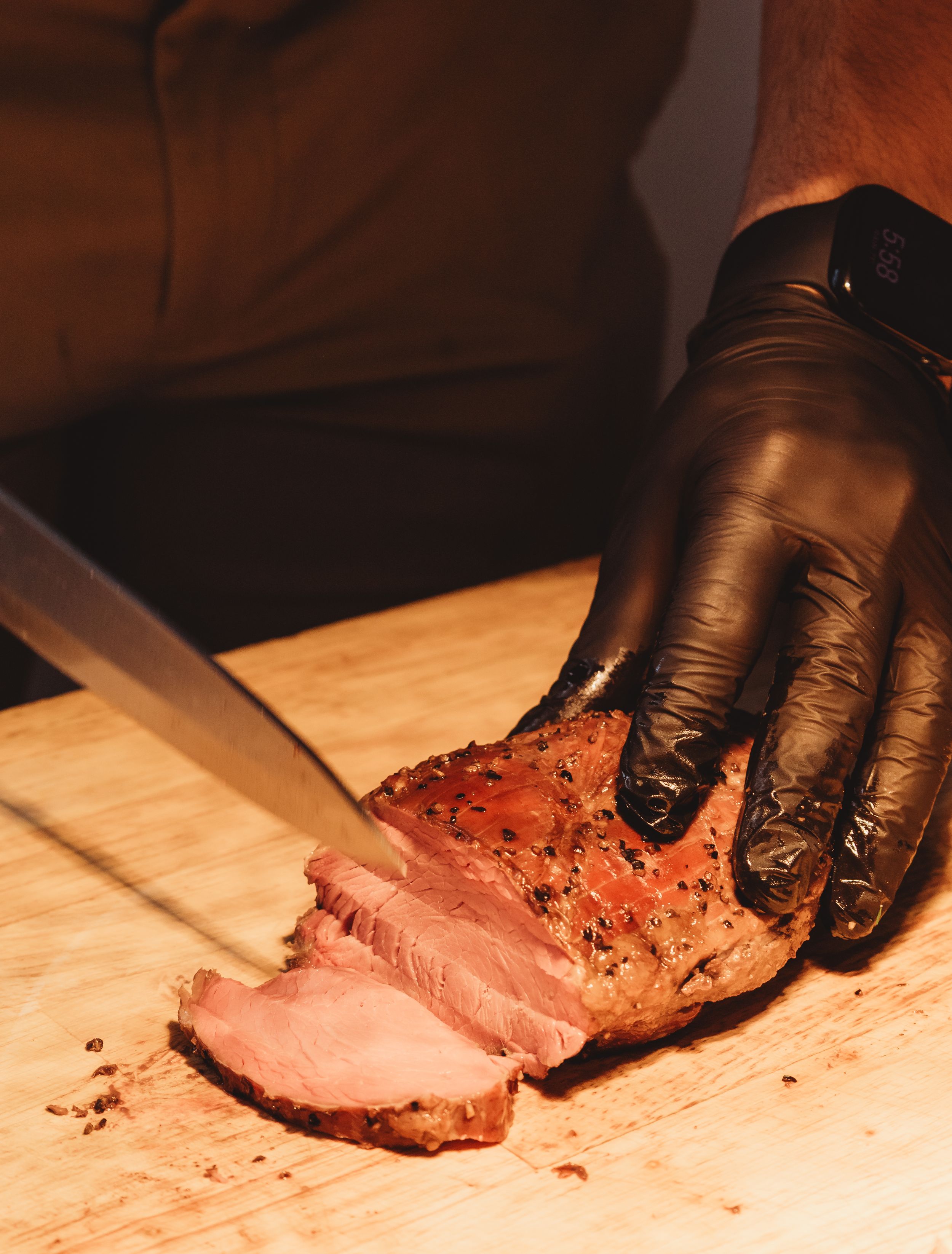 Chef in black gloves slicing seasoned roasted meat on a wooden cutting board with a sharp knife.