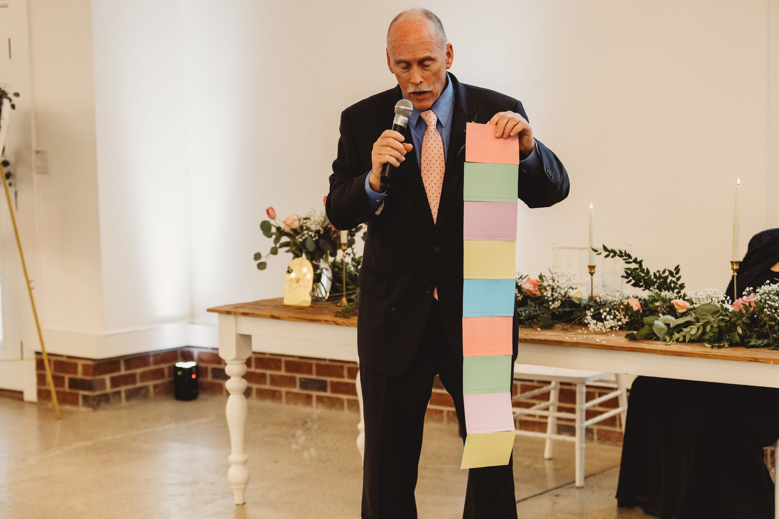 Man in black suit holding colorful stacked blocks while speaking into a microphone at an indoor event.