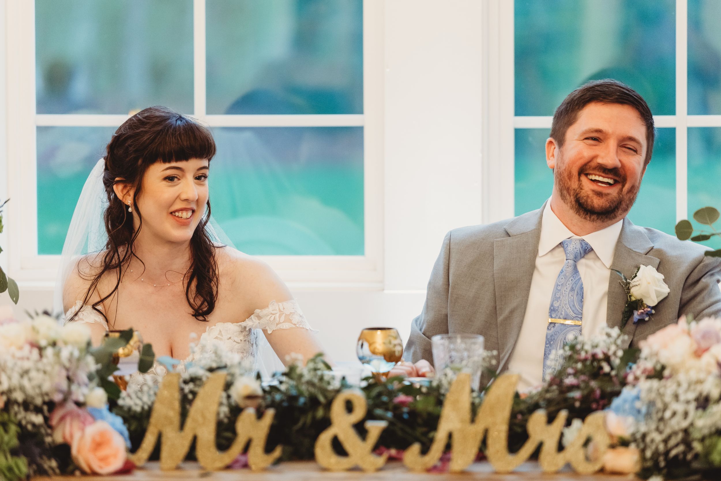 Smiling bride and groom seated at sweetheart table decorated with Mr & Mrs signs and floral arrangements.