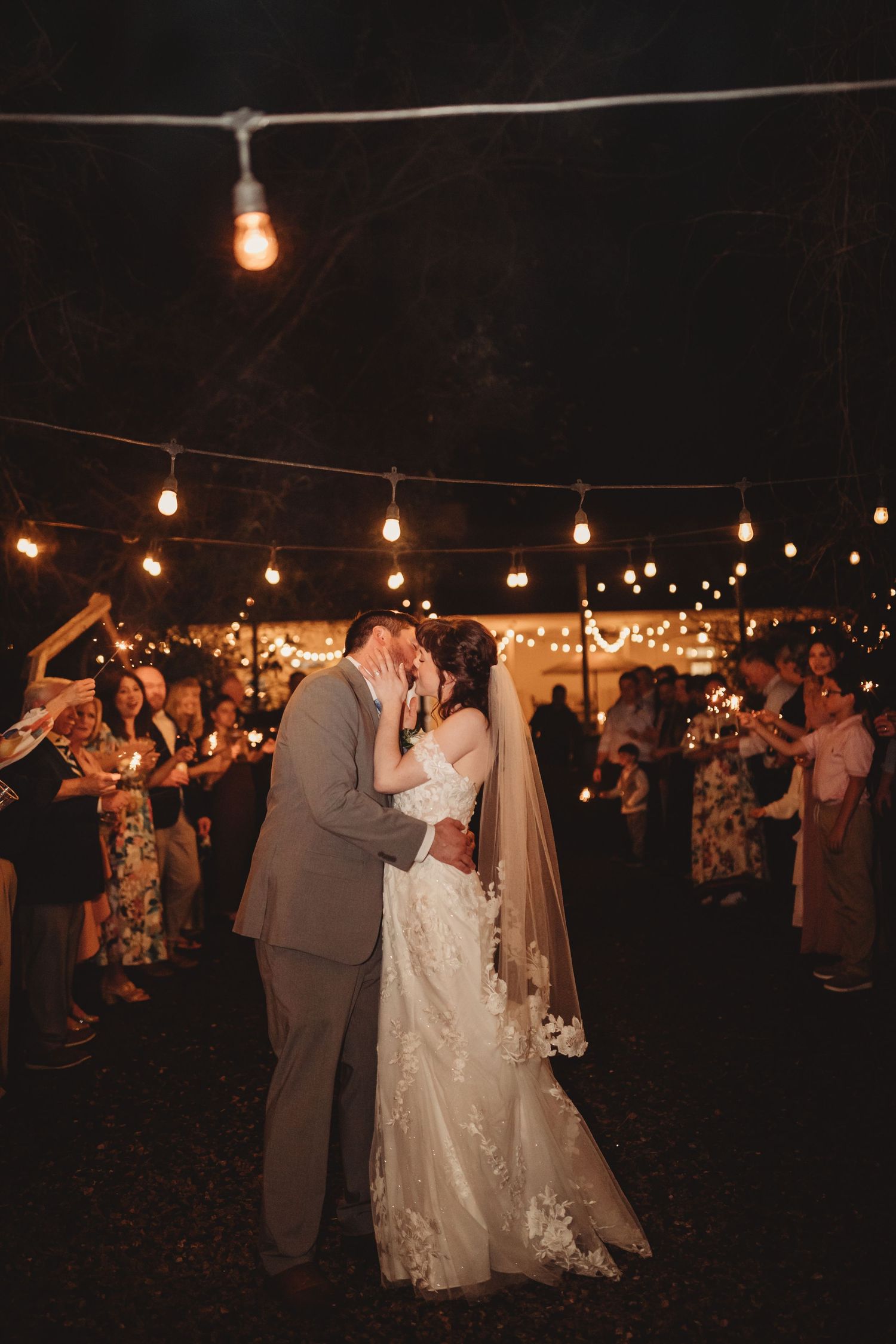 Bride and groom share a romantic kiss under warm string lights surrounded by guests holding sparklers at night.