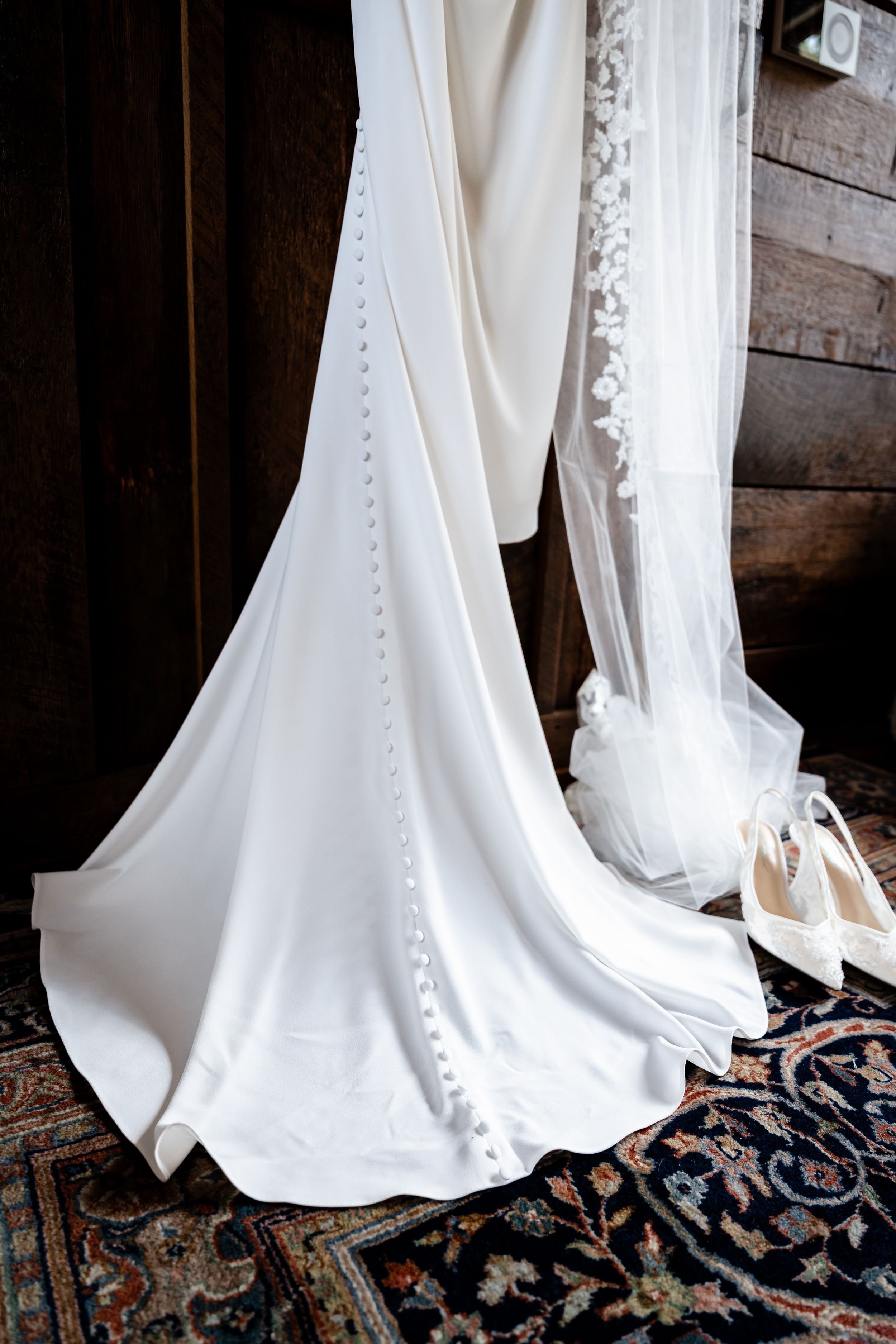 Elegant white wedding dress with button detail and lace veil hanging in a rustic wooden room with heels nearby.