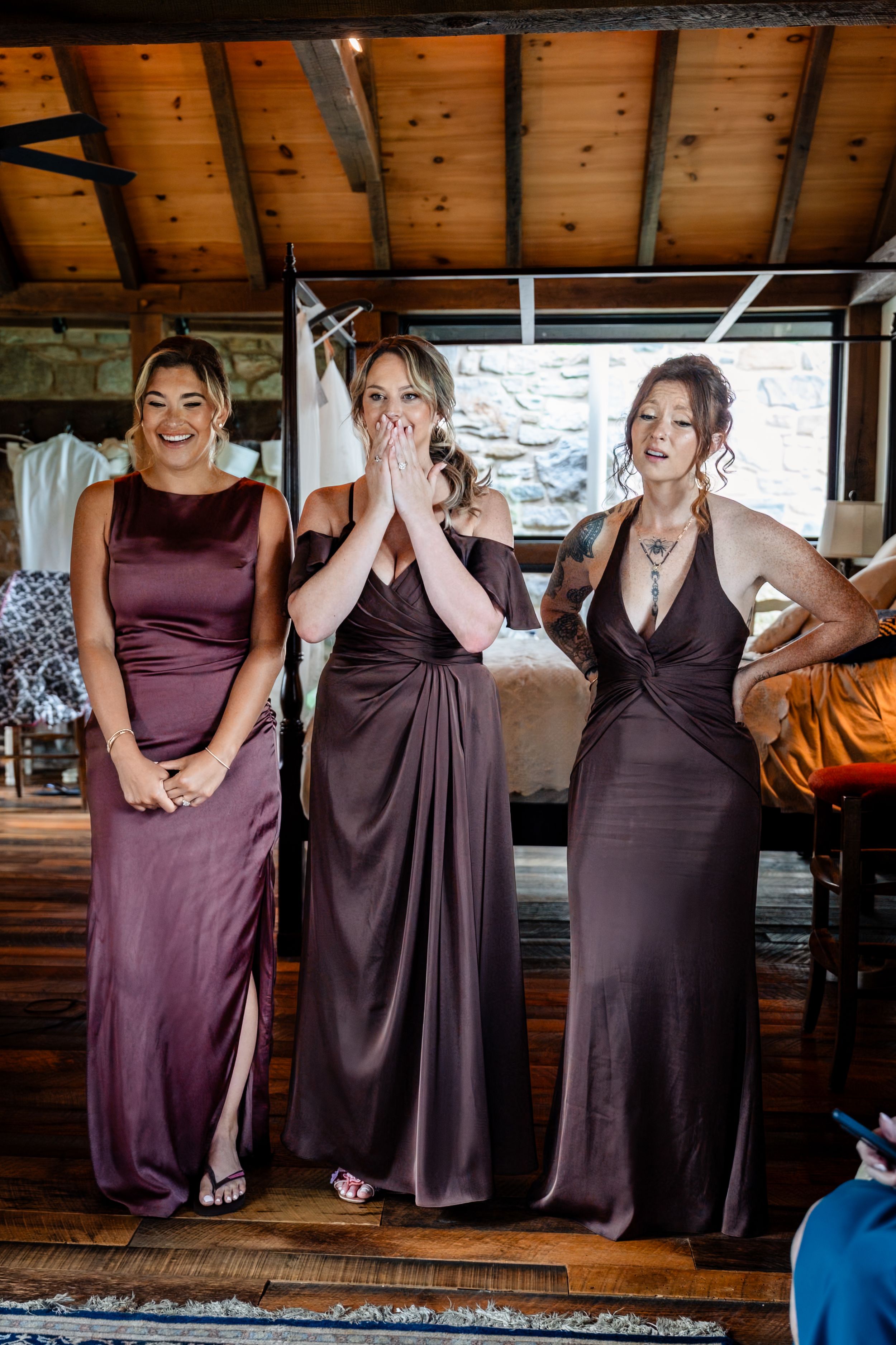 Three bridesmaids in purple satin gowns reacting with joy and surprise in a rustic wooden venue.