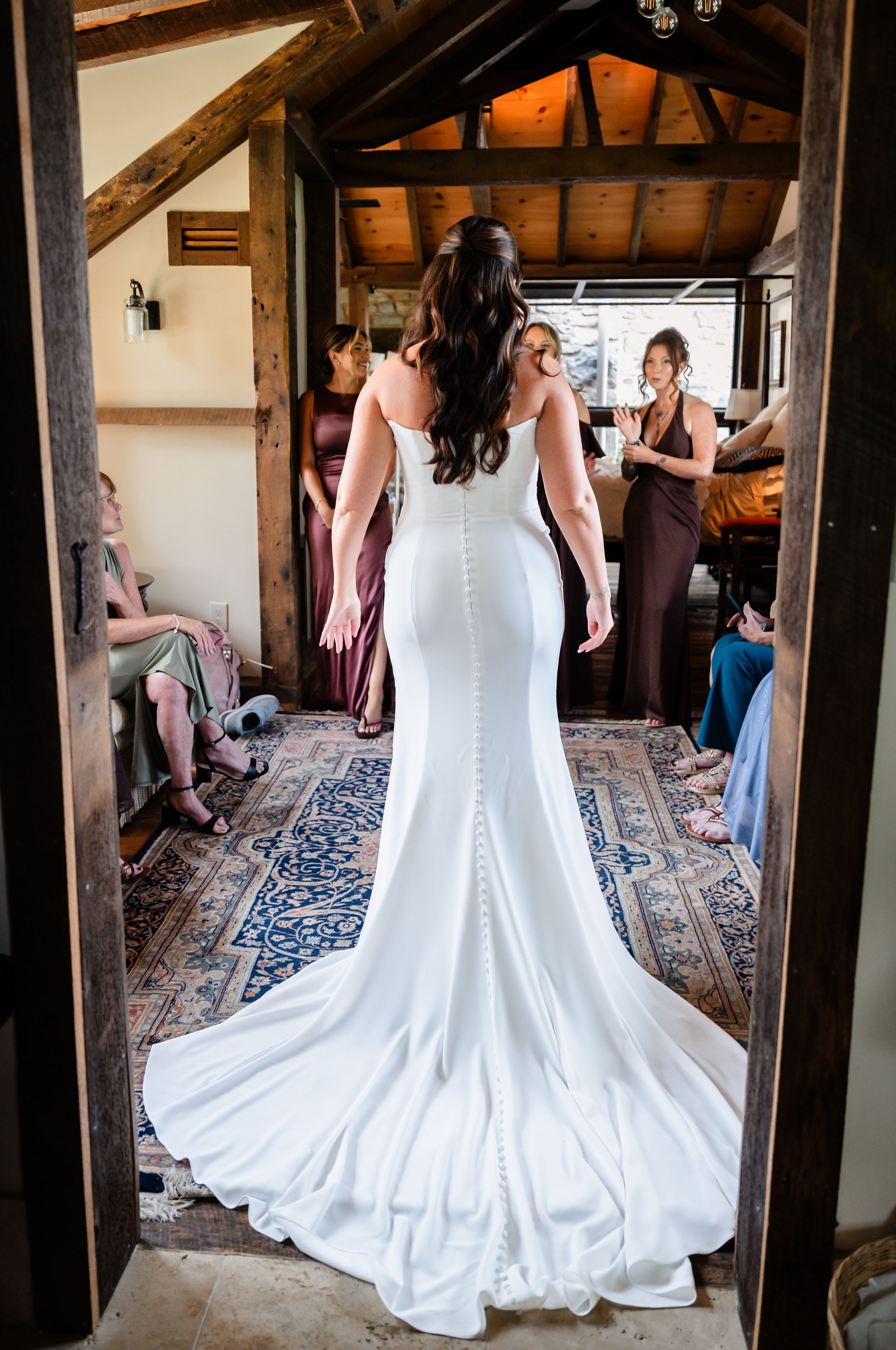 Bride in fitted white wedding gown with long train stands in rustic wooden doorway surrounded by bridesmaids.
