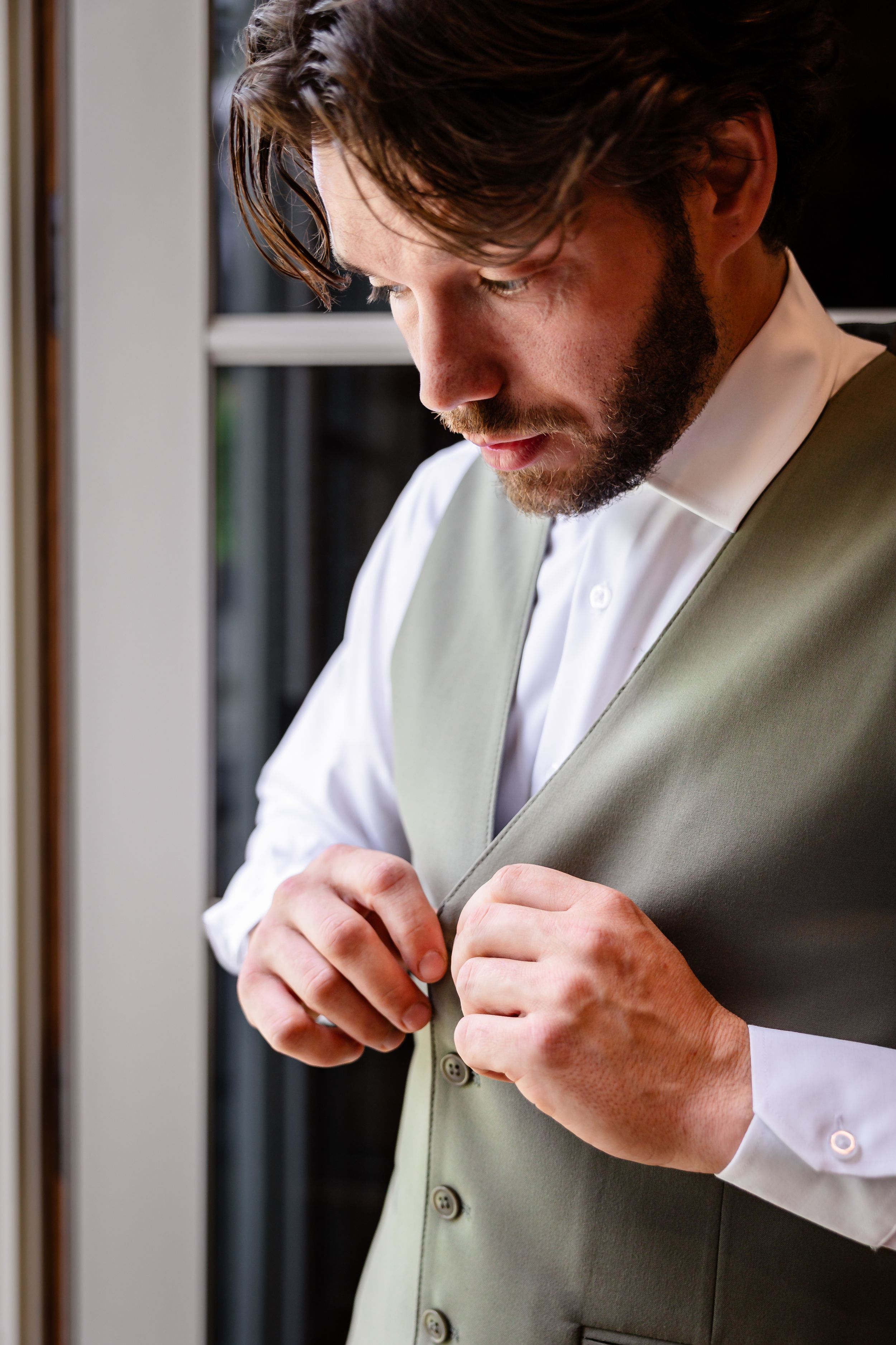 Bearded groom in white shirt and sage green vest buttons up while getting ready for wedding.