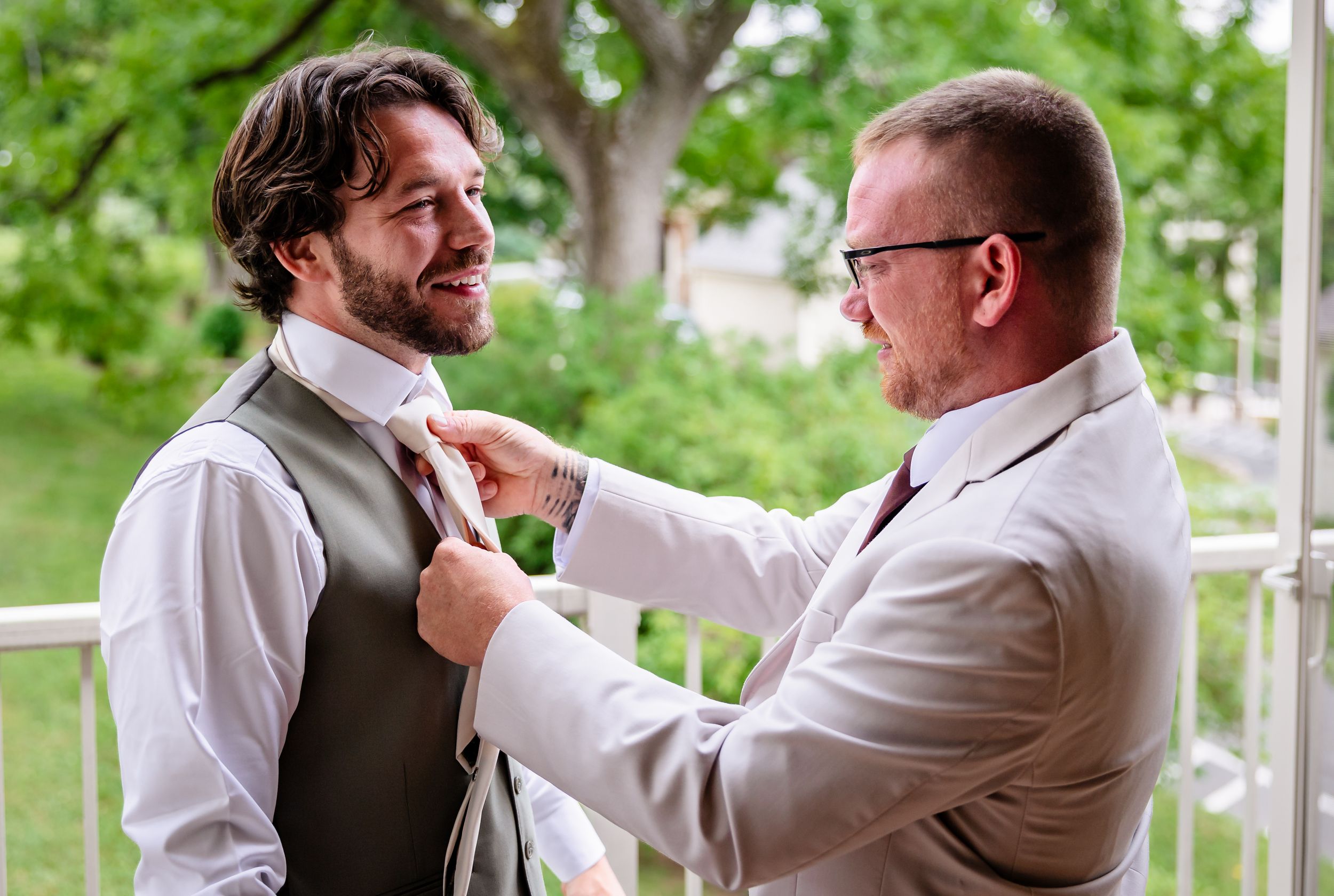 Groom smiles as groomsman adjusts his tie outdoors before wedding ceremony.