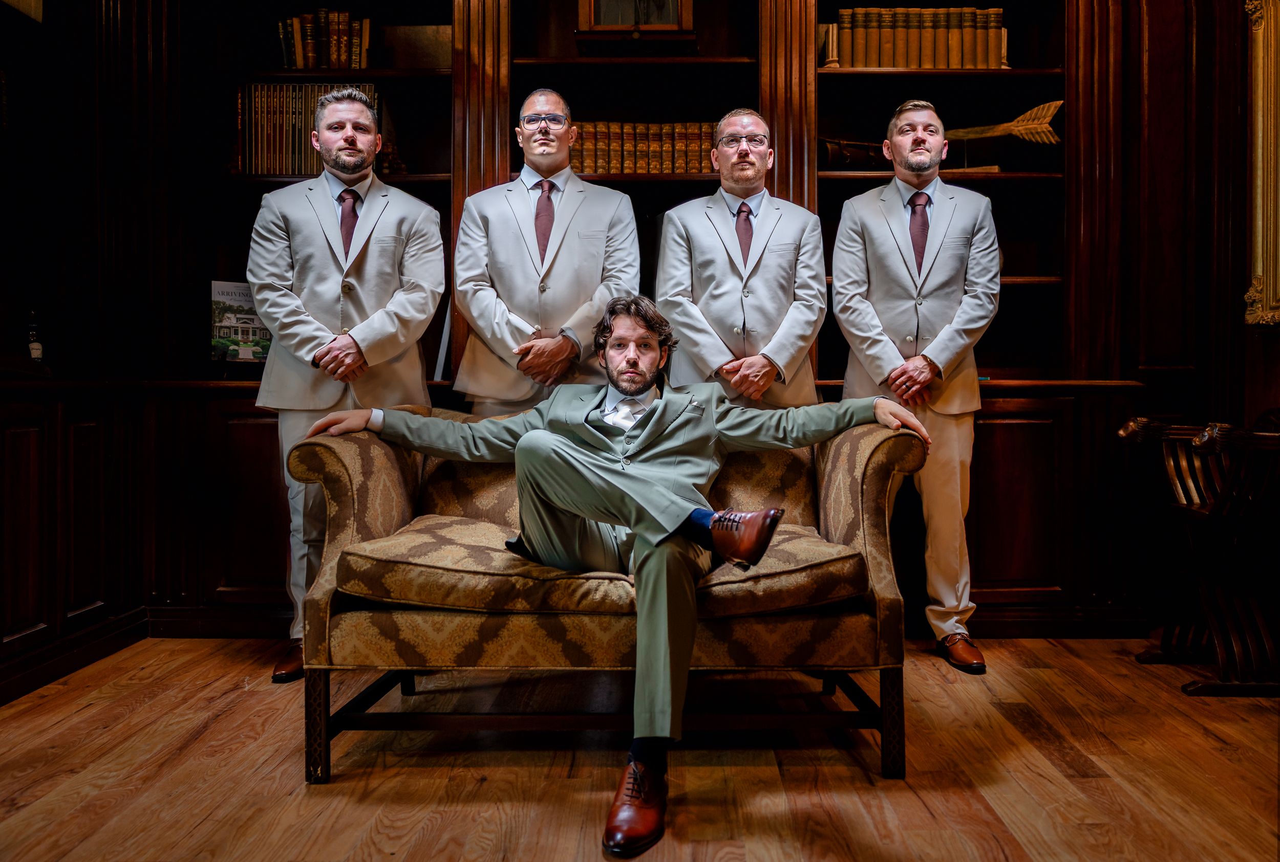Groom seated in armchair with four groomsmen standing behind in matching tan suits in library.