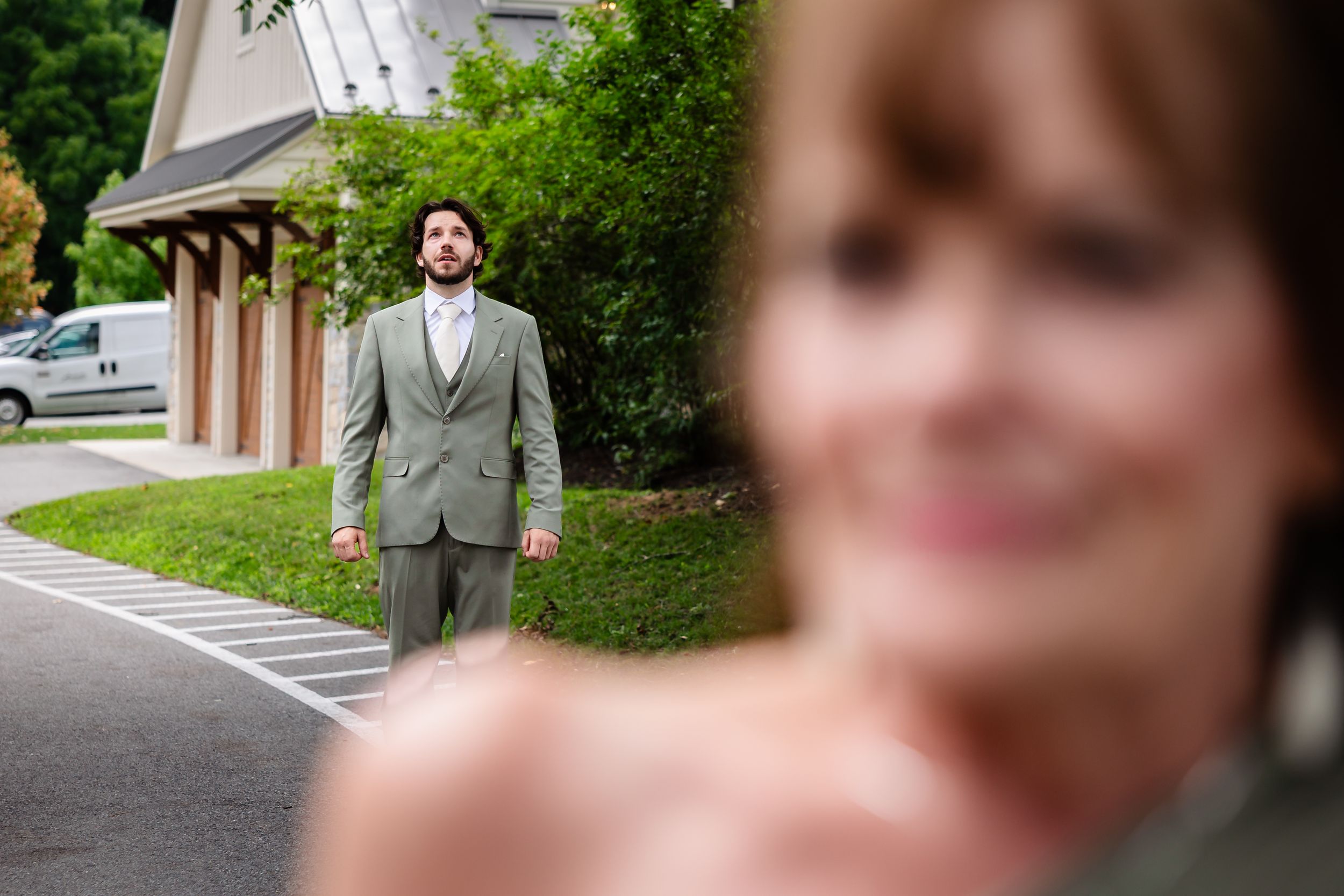 Groom in grey suit stands outdoors as blurred bride appears in foreground during wedding first look moment.