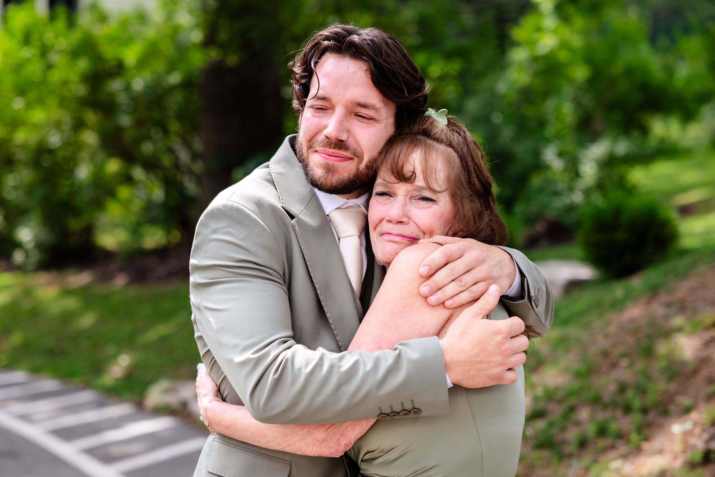 Groom in sage suit shares warm embrace with emotional family member outdoors at wedding.