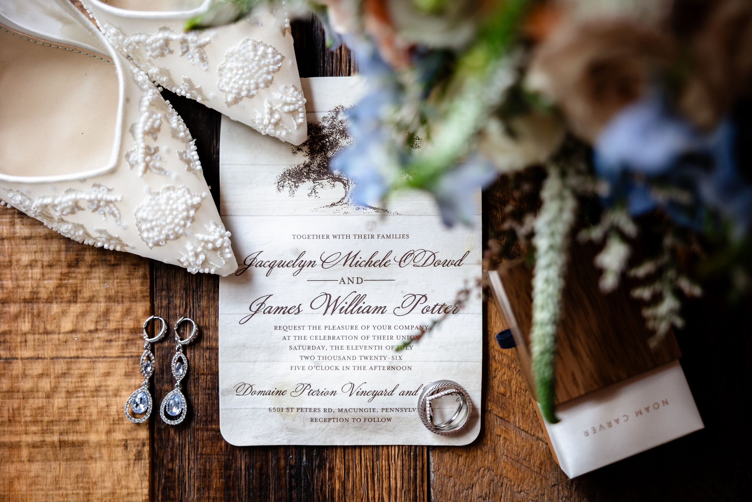 Elegant wedding invitation flat lay with floral bouquet, diamond earrings, and ring on rustic wood table.