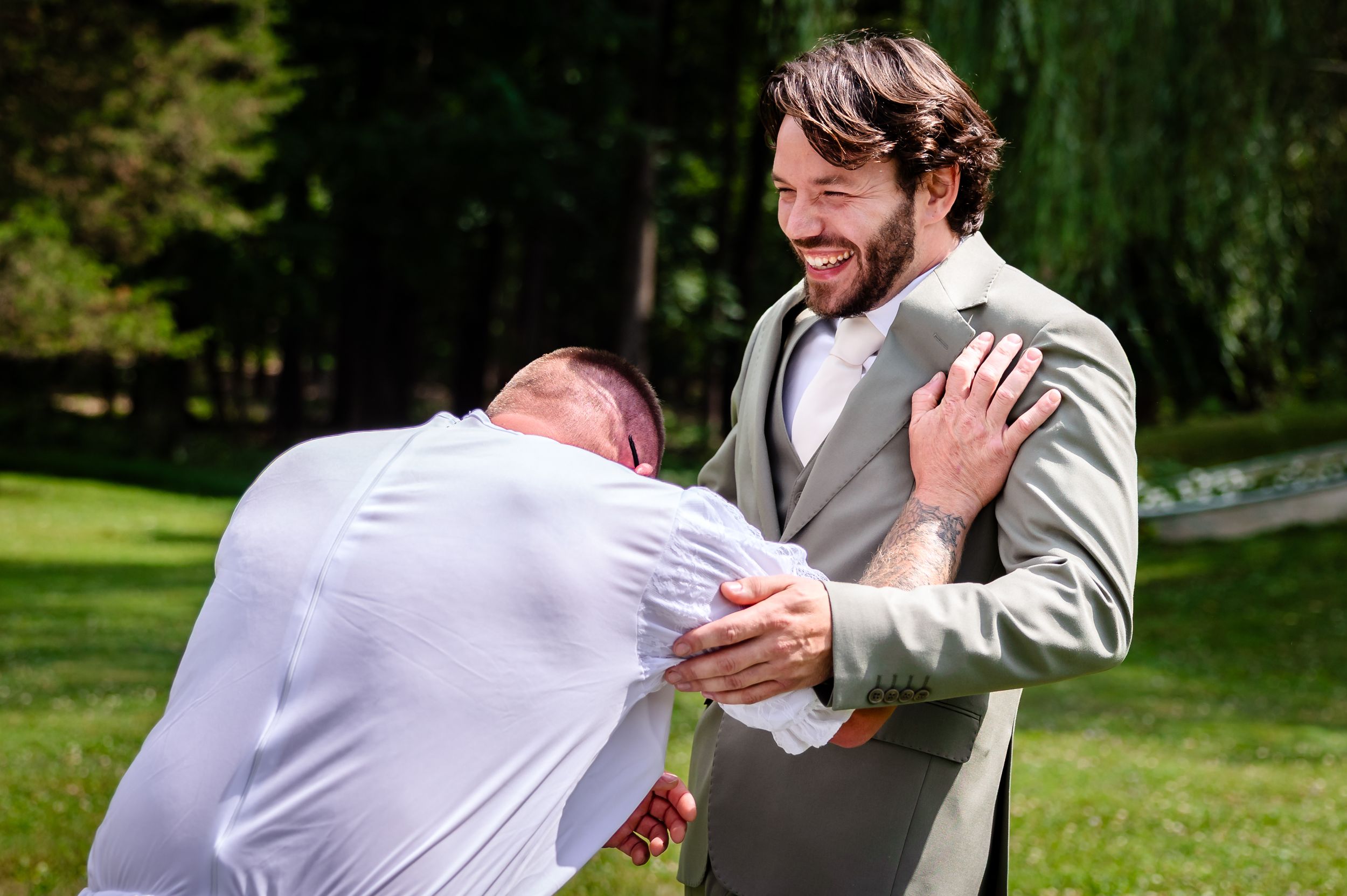 Groom laughs joyfully as a groomsman bows in congratulations during an outdoor wedding celebration.