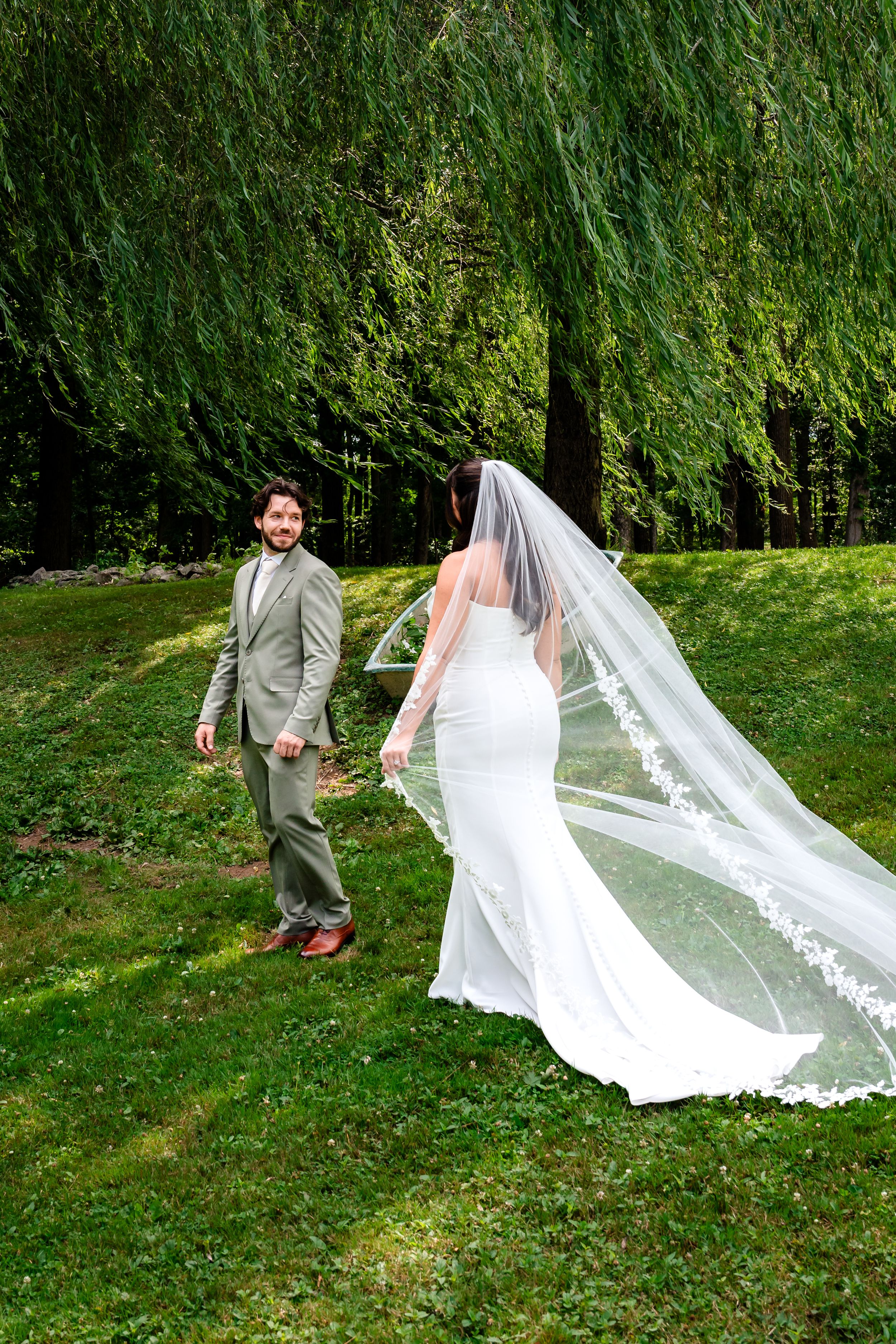 Bride in white gown with flowing lace veil walks with bearded groom in gray suit near weeping willow tree.