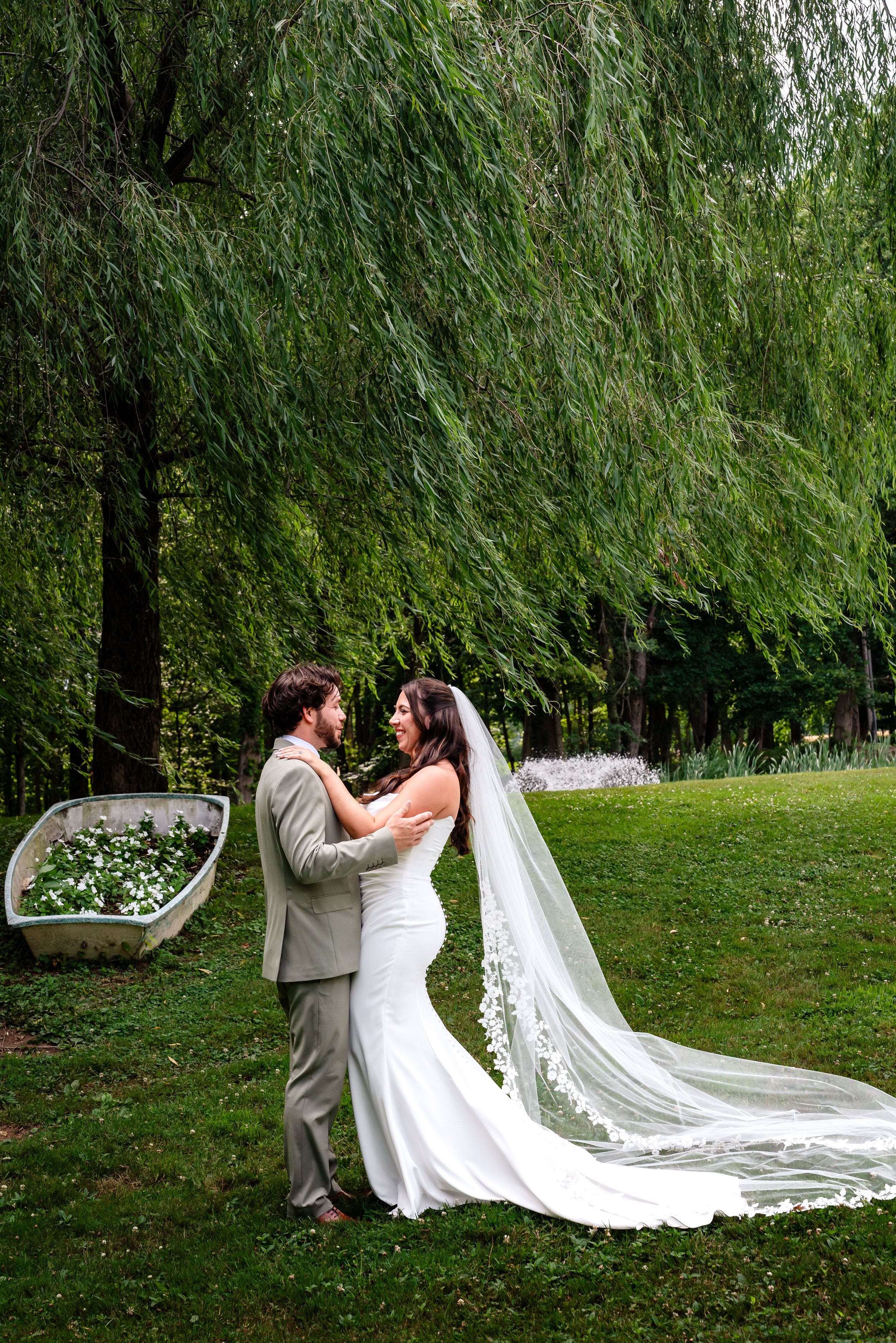 Bride and groom embrace romantically beneath a weeping willow tree with long lace veil flowing.