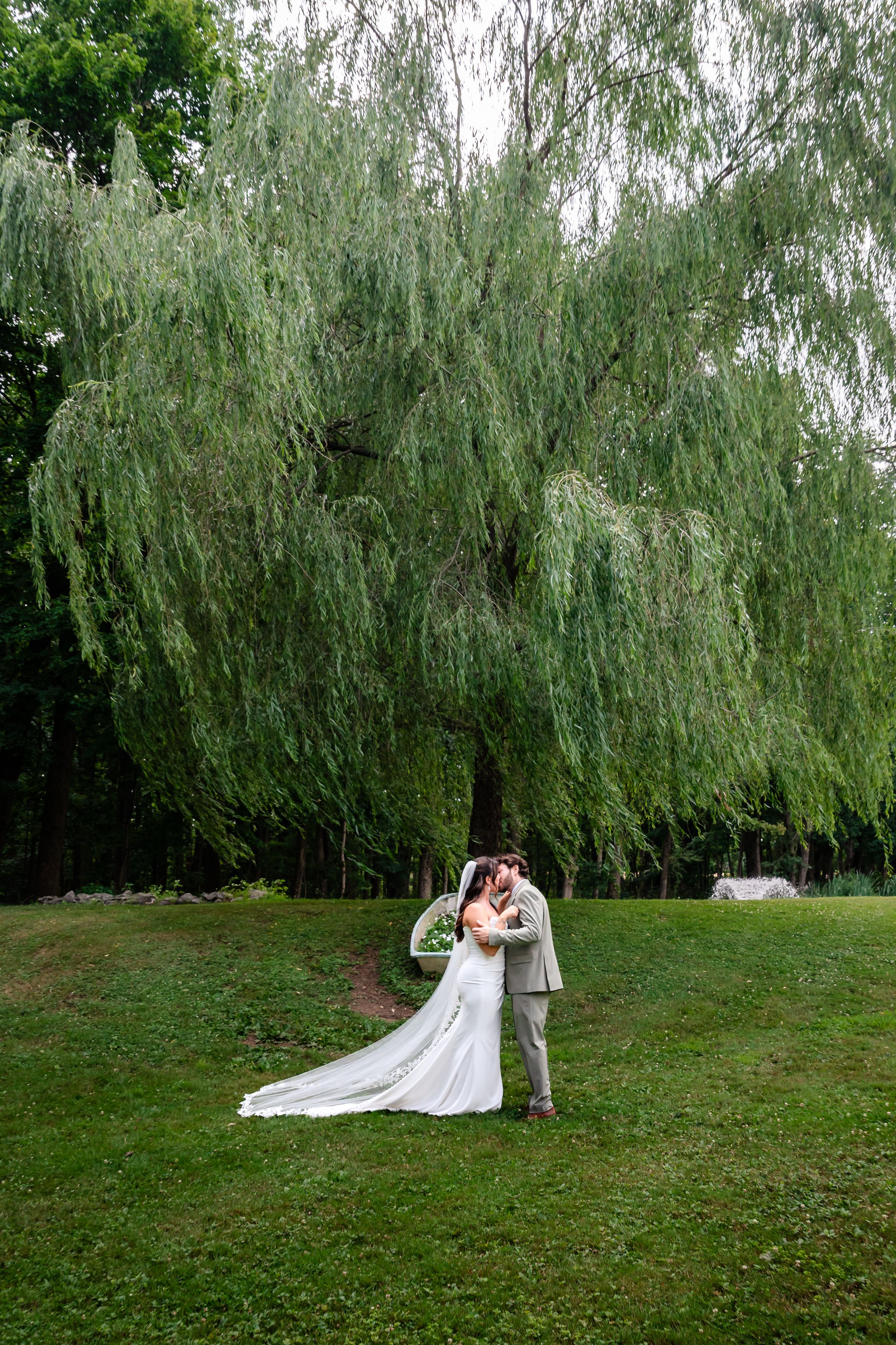 Bride and groom share a kiss under a large weeping willow tree on lush green lawn.