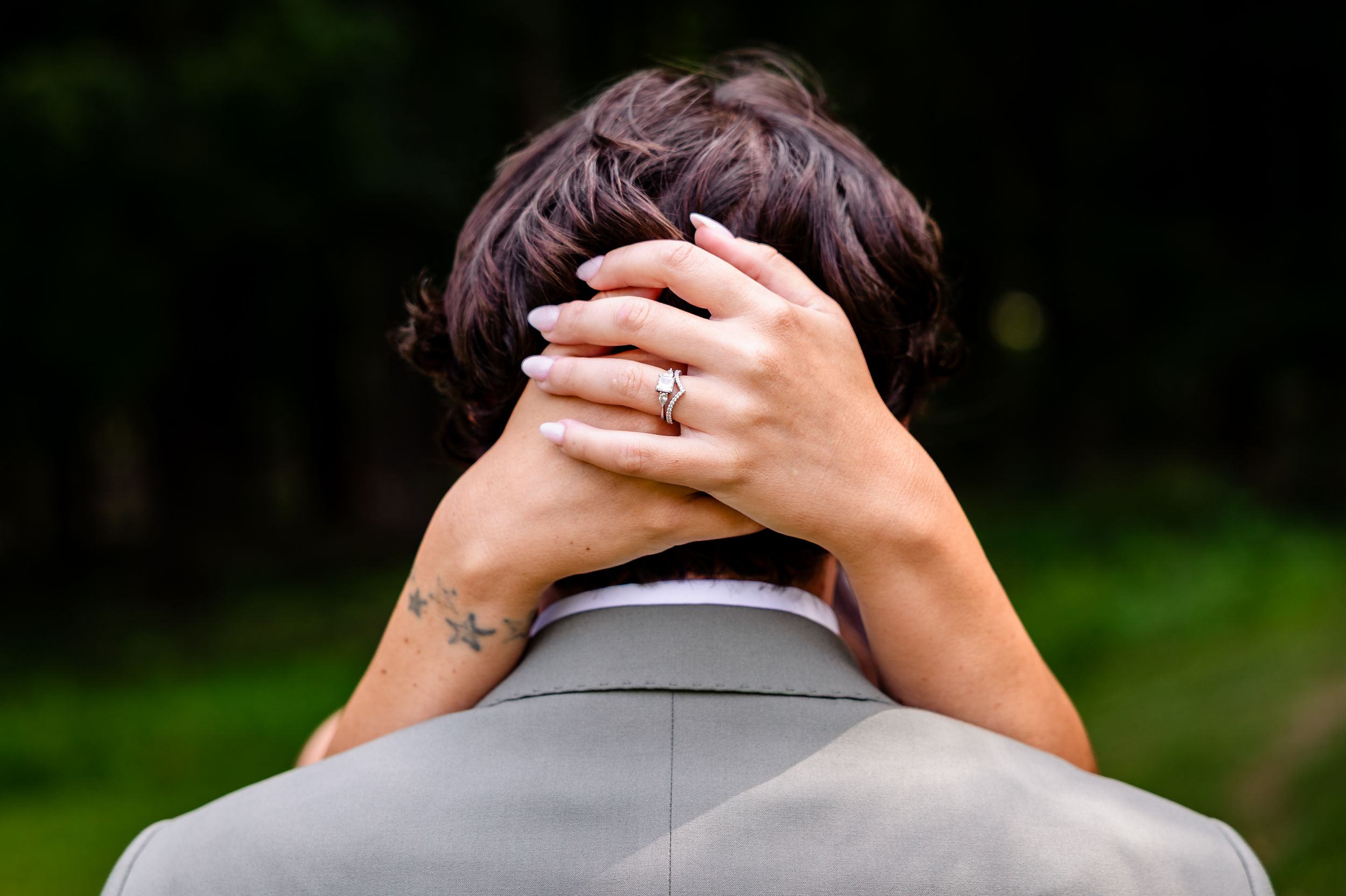 Close-up of bride's hands with engagement ring resting on groom's neck at outdoor wedding.