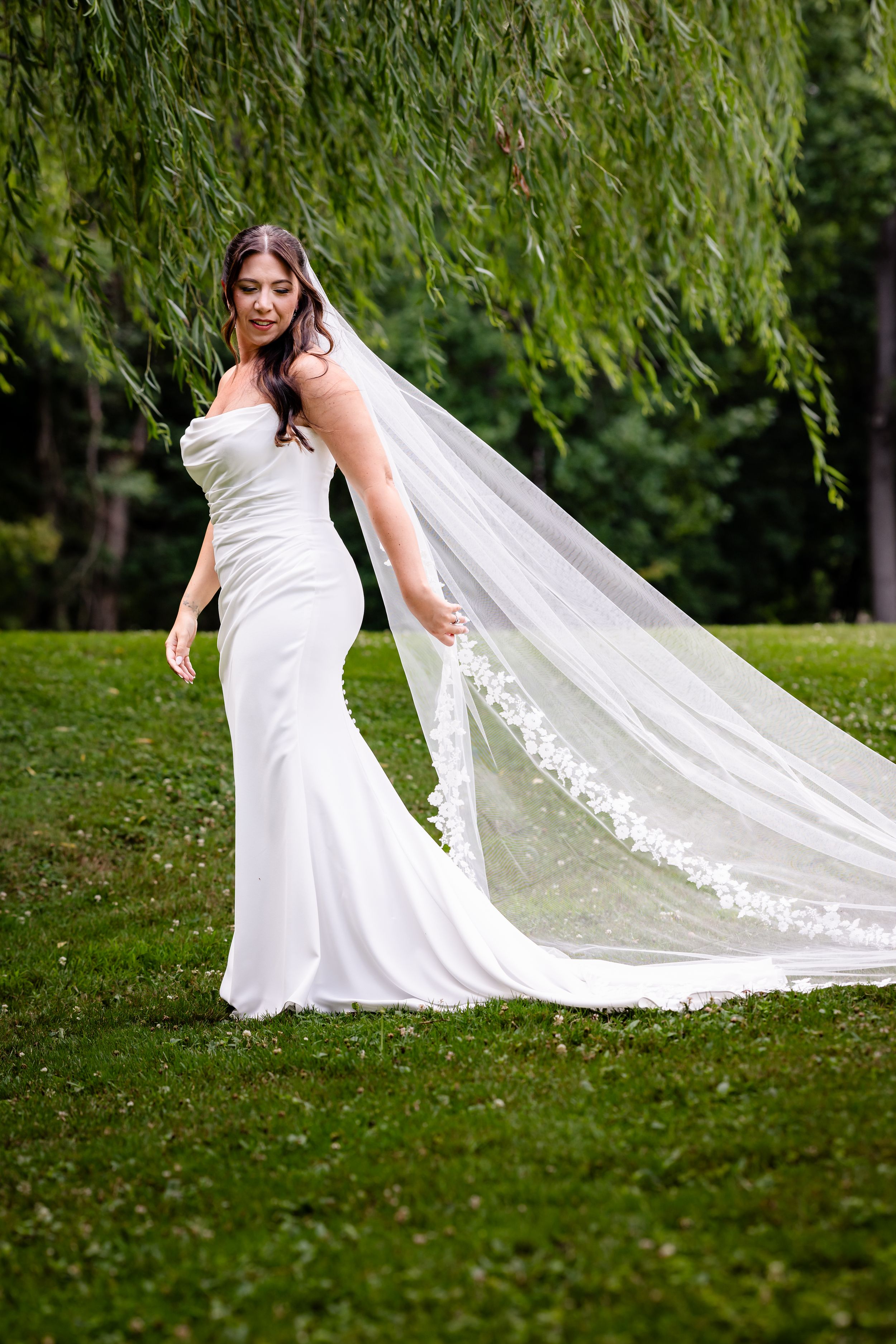 Bride in white strapless mermaid gown with long lace veil standing on green grass near weeping willow tree.