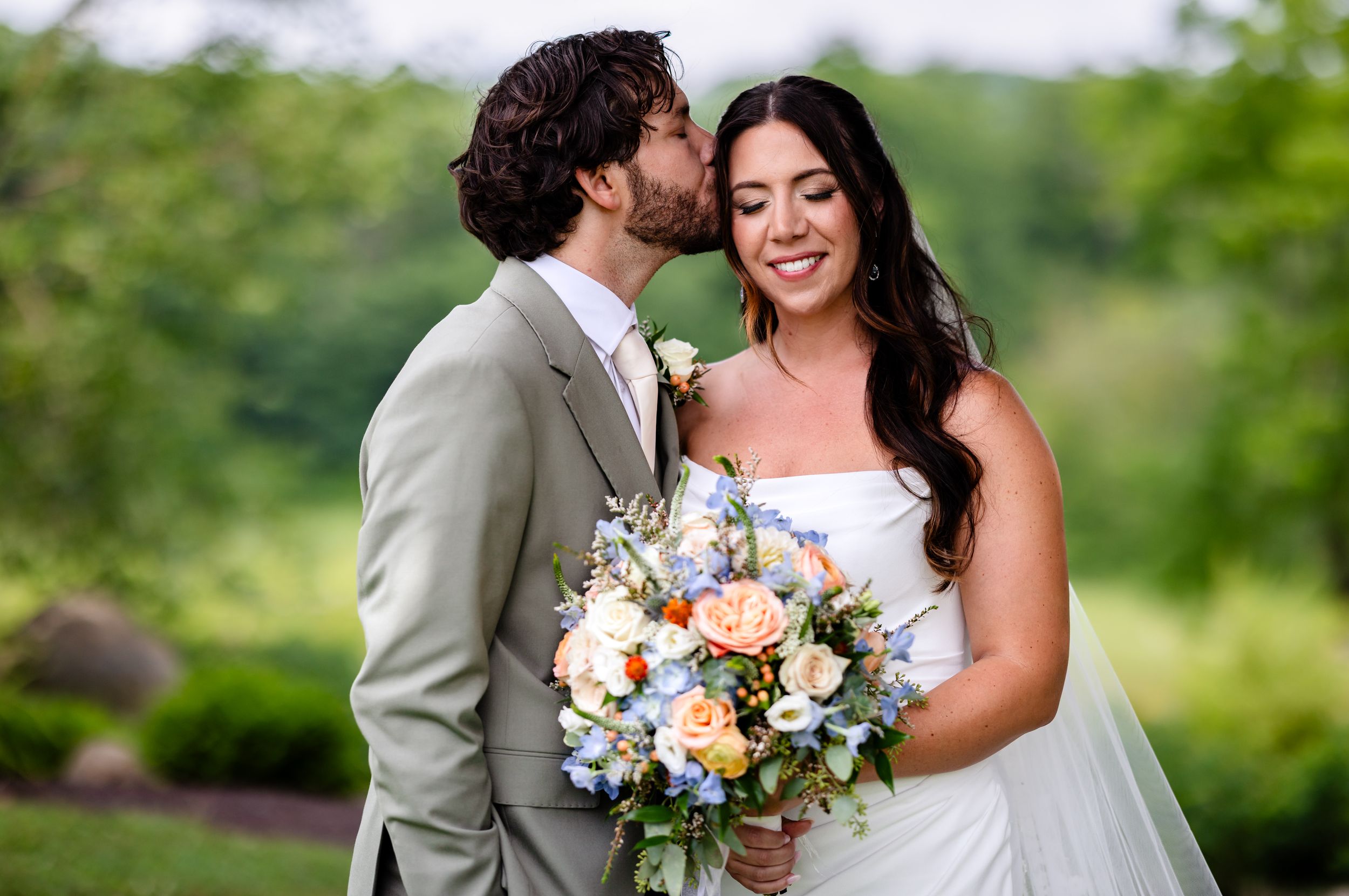 Groom in gray suit kissing bride's cheek as she smiles holding a colorful spring bouquet.