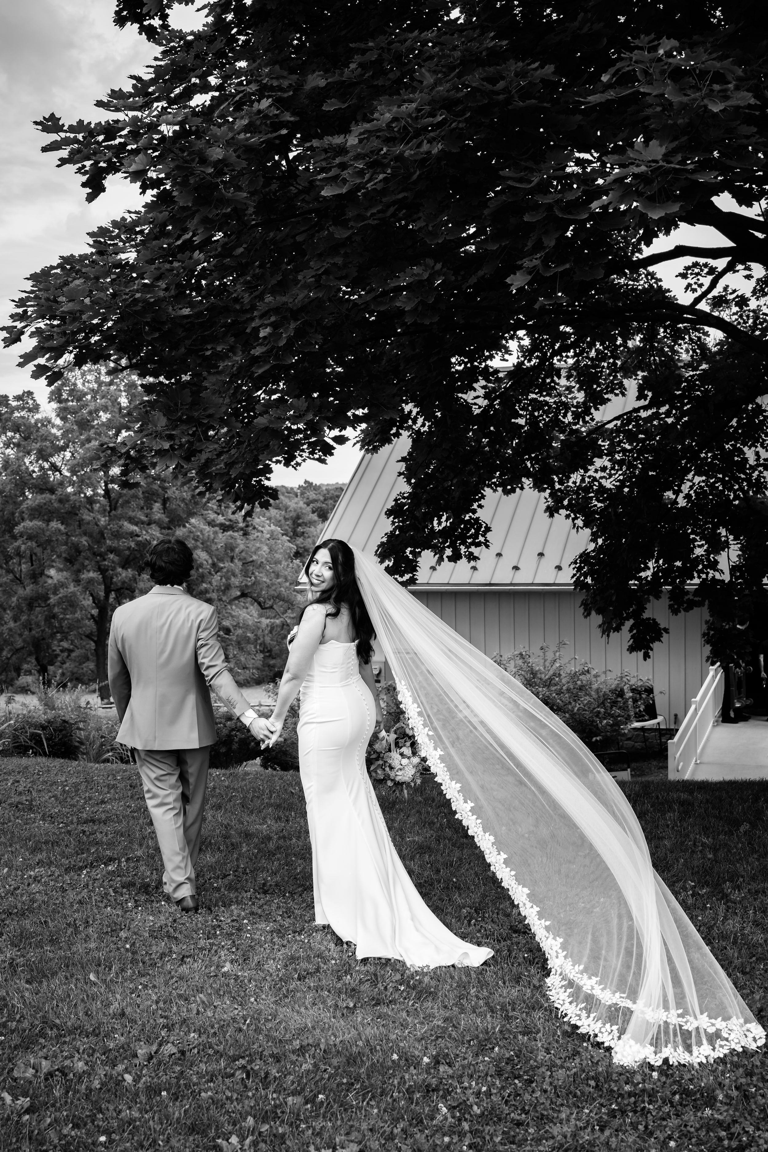 Black and white photo of bride and groom holding hands outdoors, brides lace veil flowing in the wind.