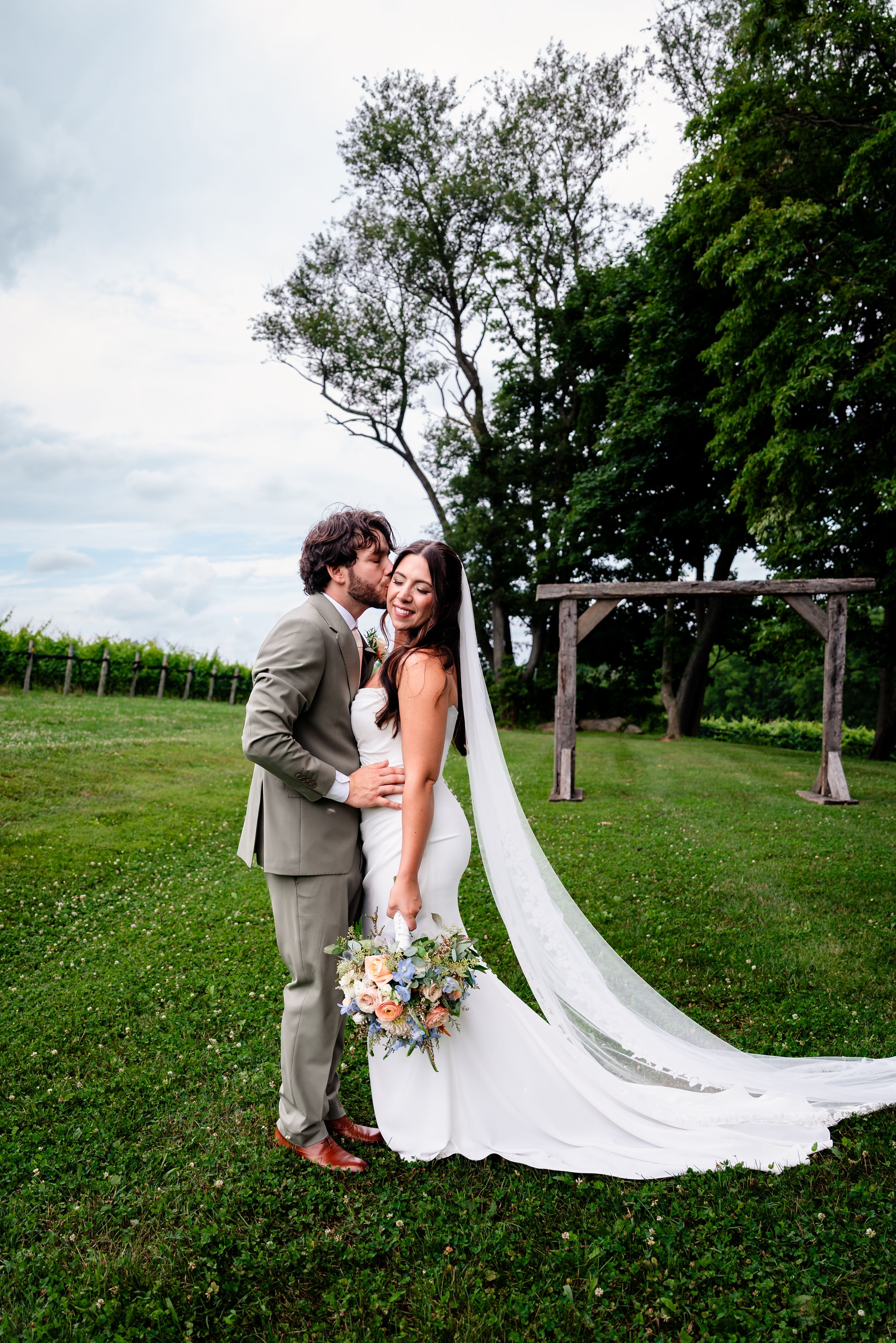Bride and groom share a romantic kiss outdoors at a vineyard wedding, with her flowing veil trailing on green grass.