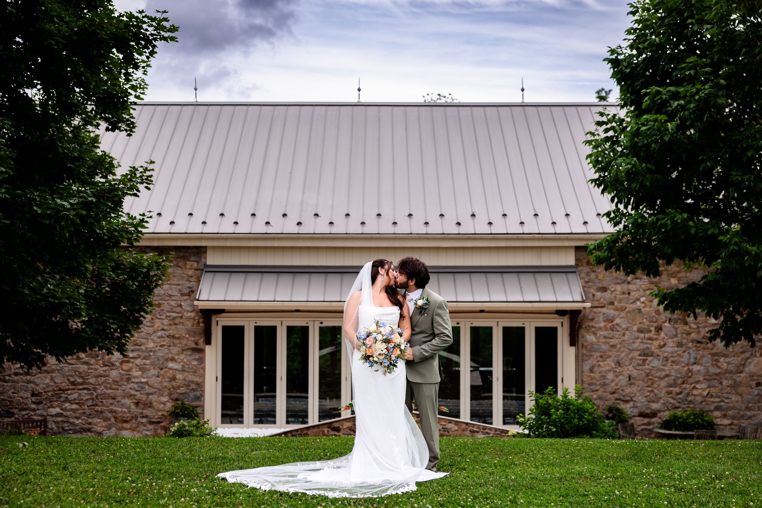 Bride and groom share a romantic kiss outside a stone building with a metal roof on their wedding day.