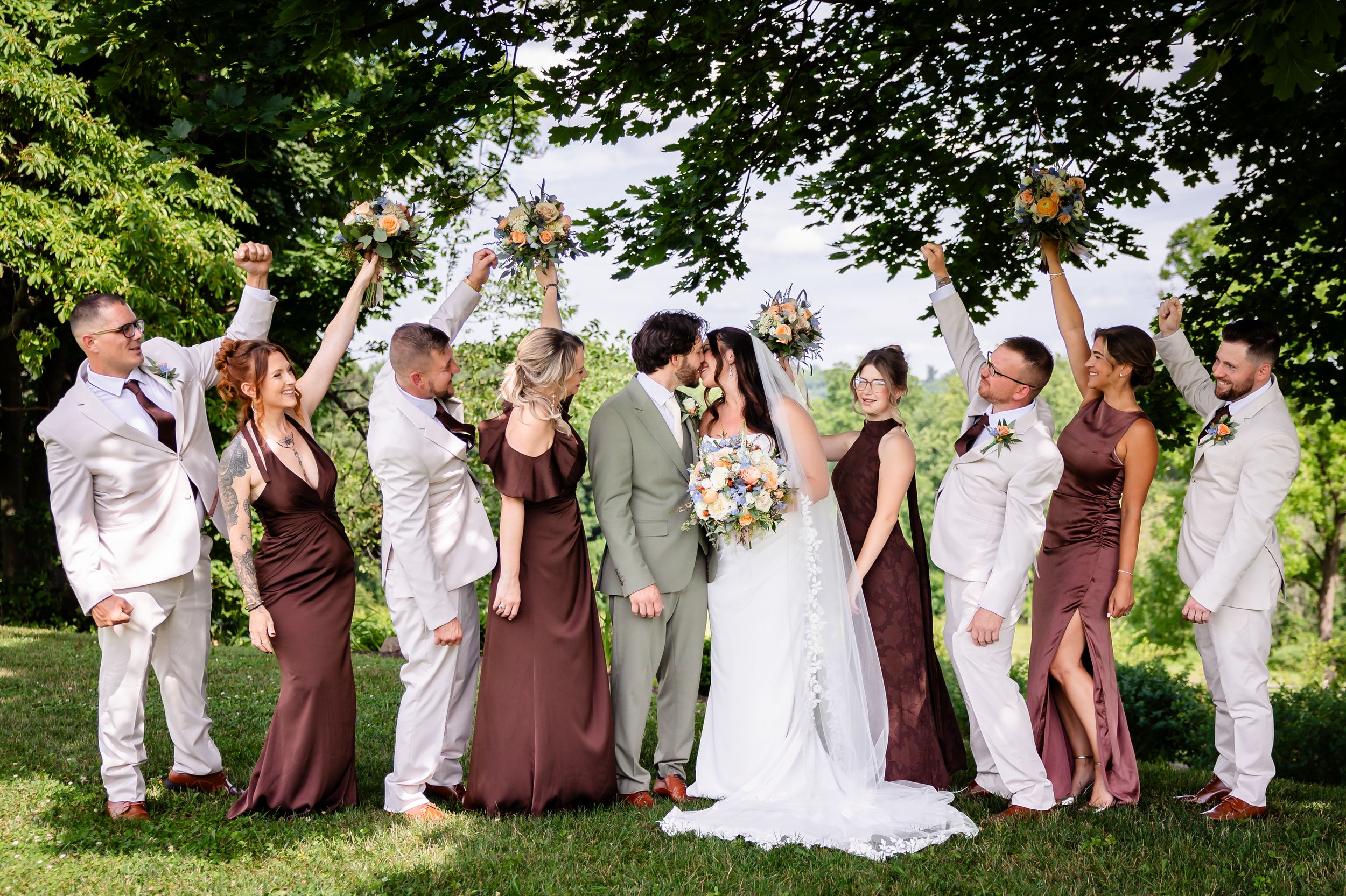 Wedding party celebrating outdoors, bridesmaids in brown dresses, groomsmen in white suits, bride and groom kissing center.