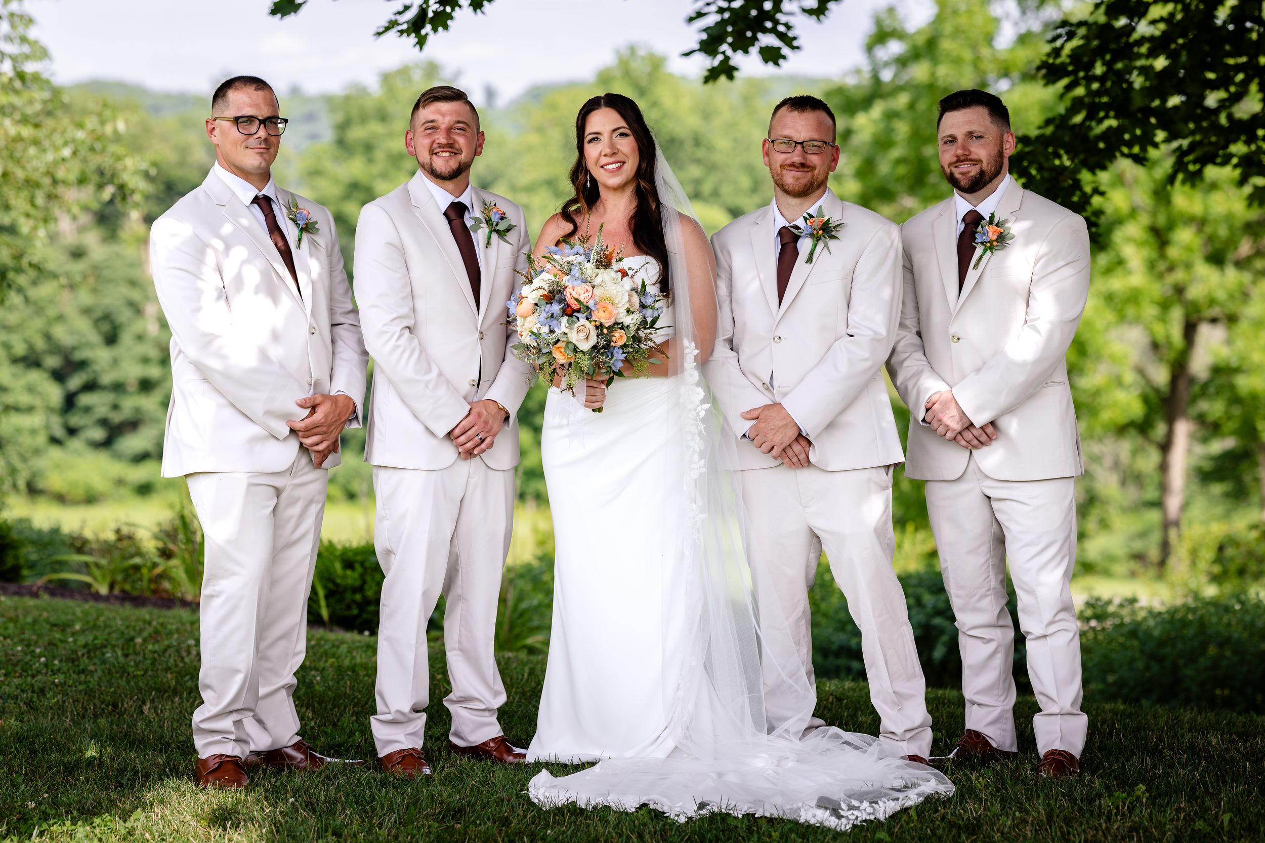 Bride in white gown poses with four groomsmen in white suits outdoors at a wedding.