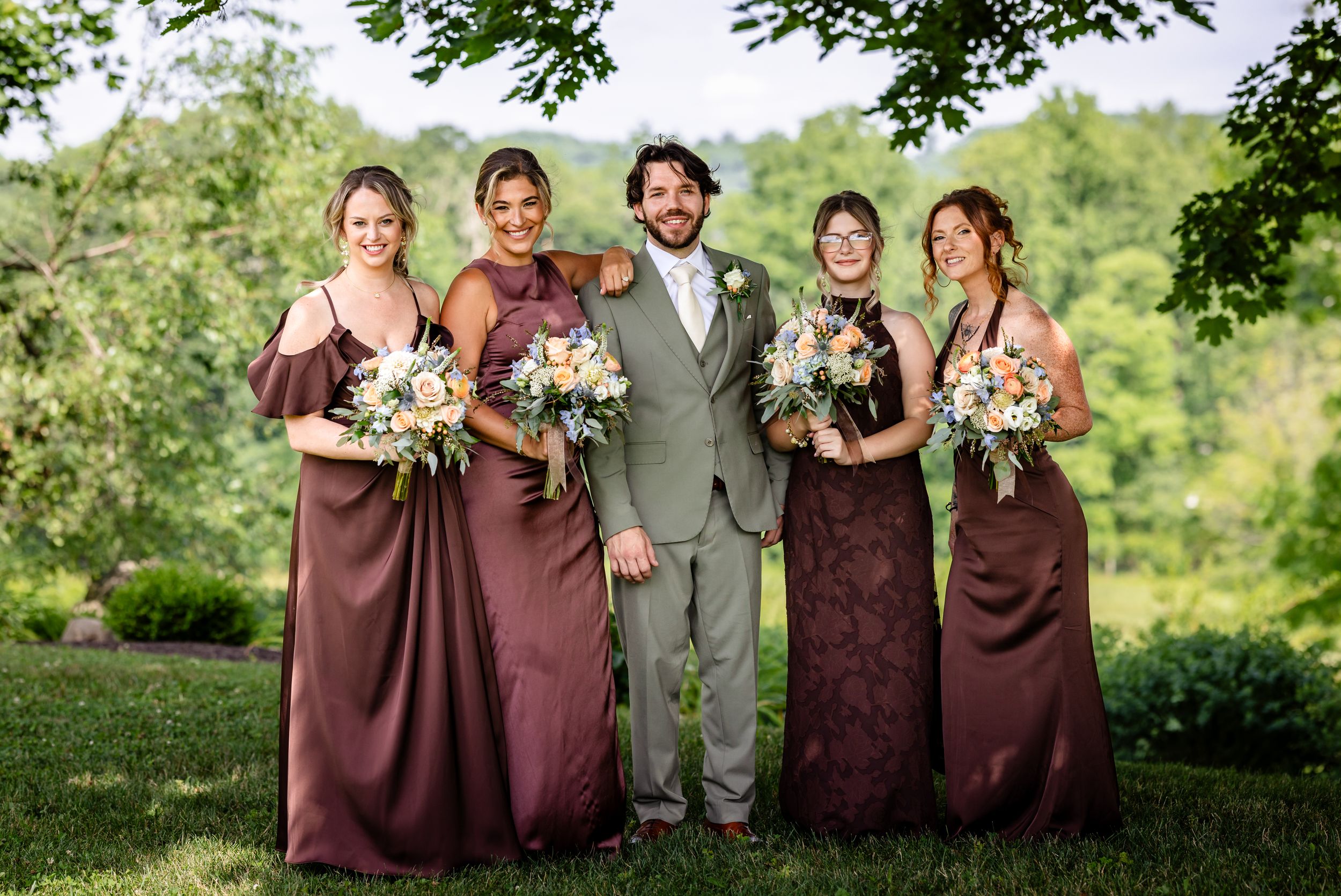 Groom in gray suit poses with four bridesmaids in mauve dresses holding floral bouquets outdoors.