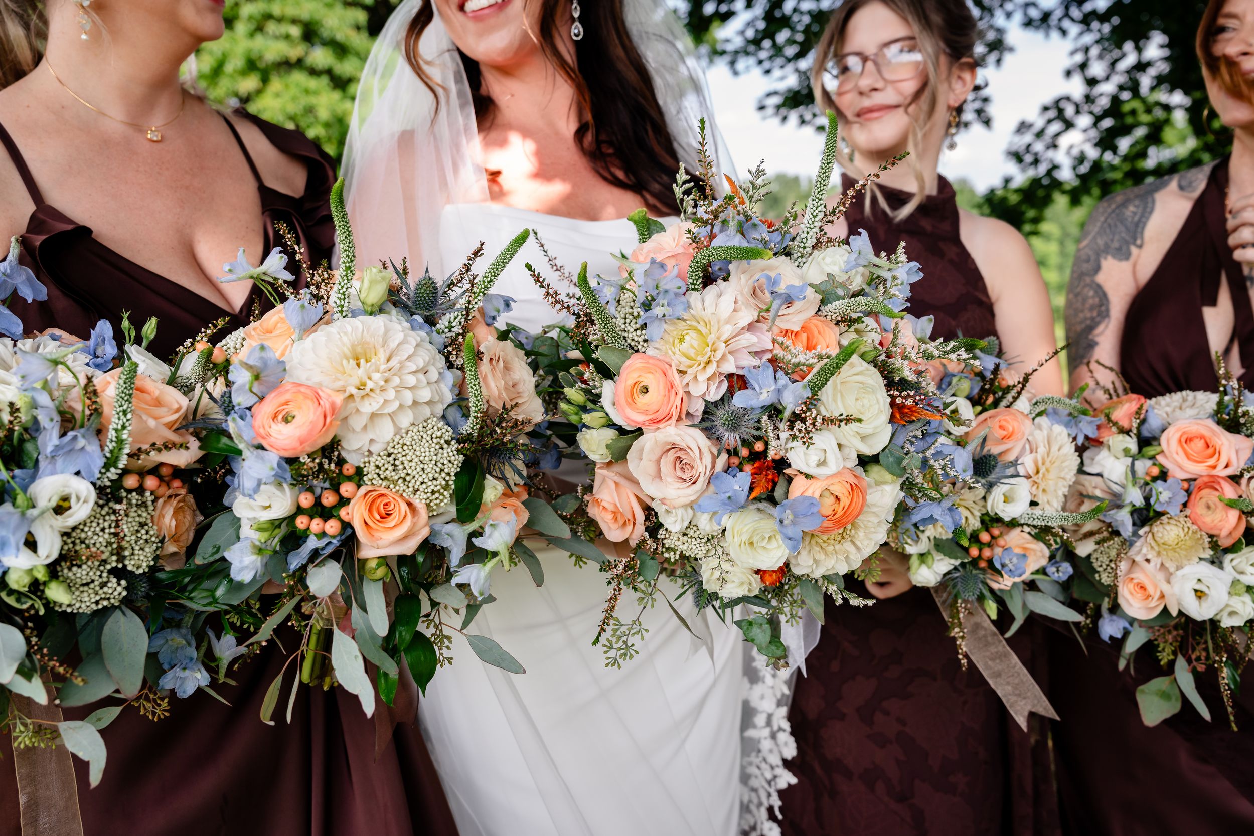 Bride and bridesmaids in burgundy dresses holding lush bouquets of peach roses, white flowers, and blue blooms outdoors.