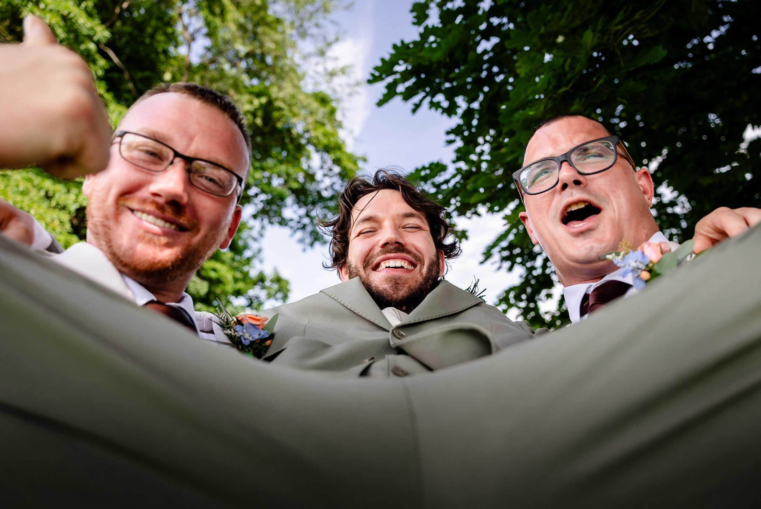 Three smiling men in formal wedding attire photographed from below against a blue sky and green trees.