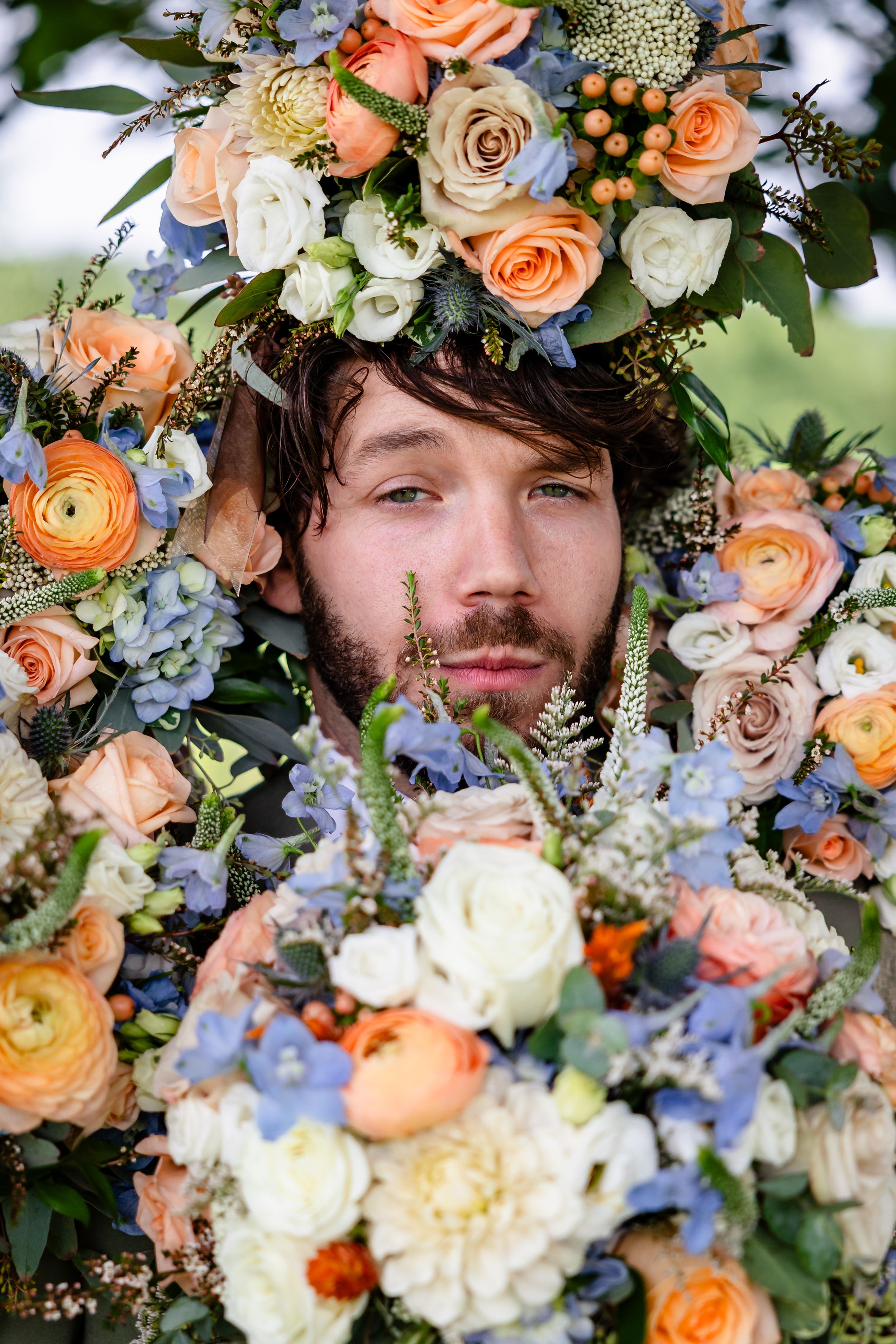 Man with blue eyes surrounded by colorful floral arrangement featuring peach roses, white blooms, and blue flowers.