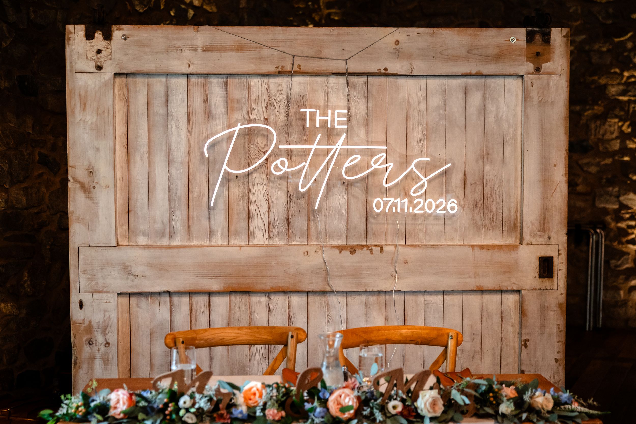 Rustic wooden backdrop with neon sign reading The Potters 07.11.2026 above sweetheart table with floral arrangements.