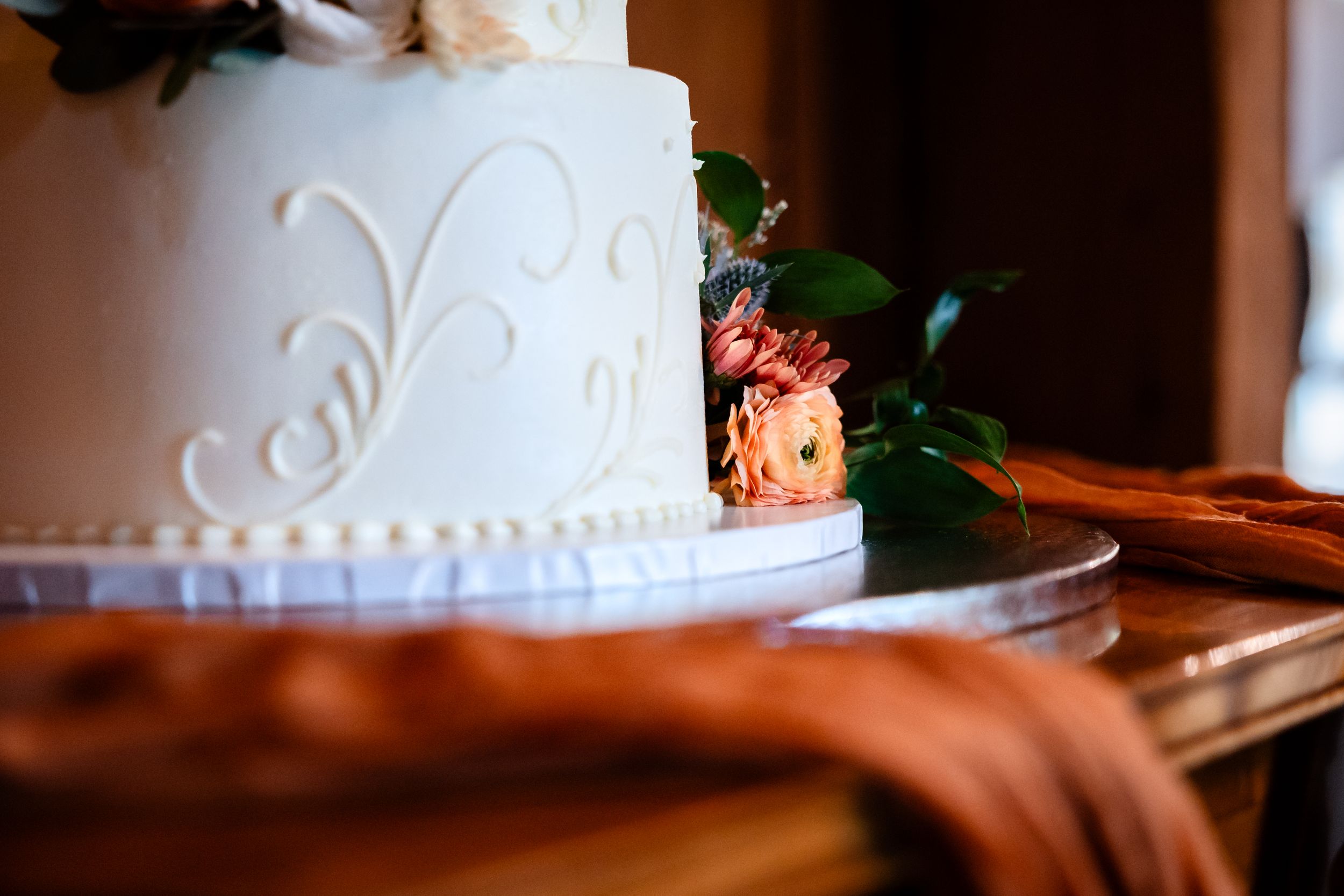 Elegant two-tier white wedding cake decorated with floral swirls and fresh peach roses on wooden table.
