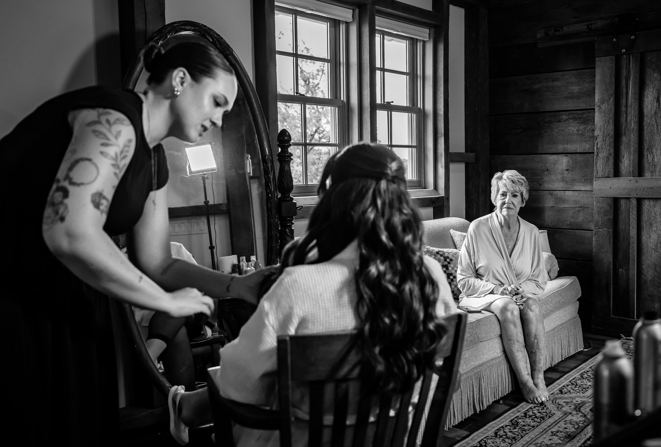 Black and white photo of a bride getting ready with a stylist and smiling woman in a robe in a rustic room.