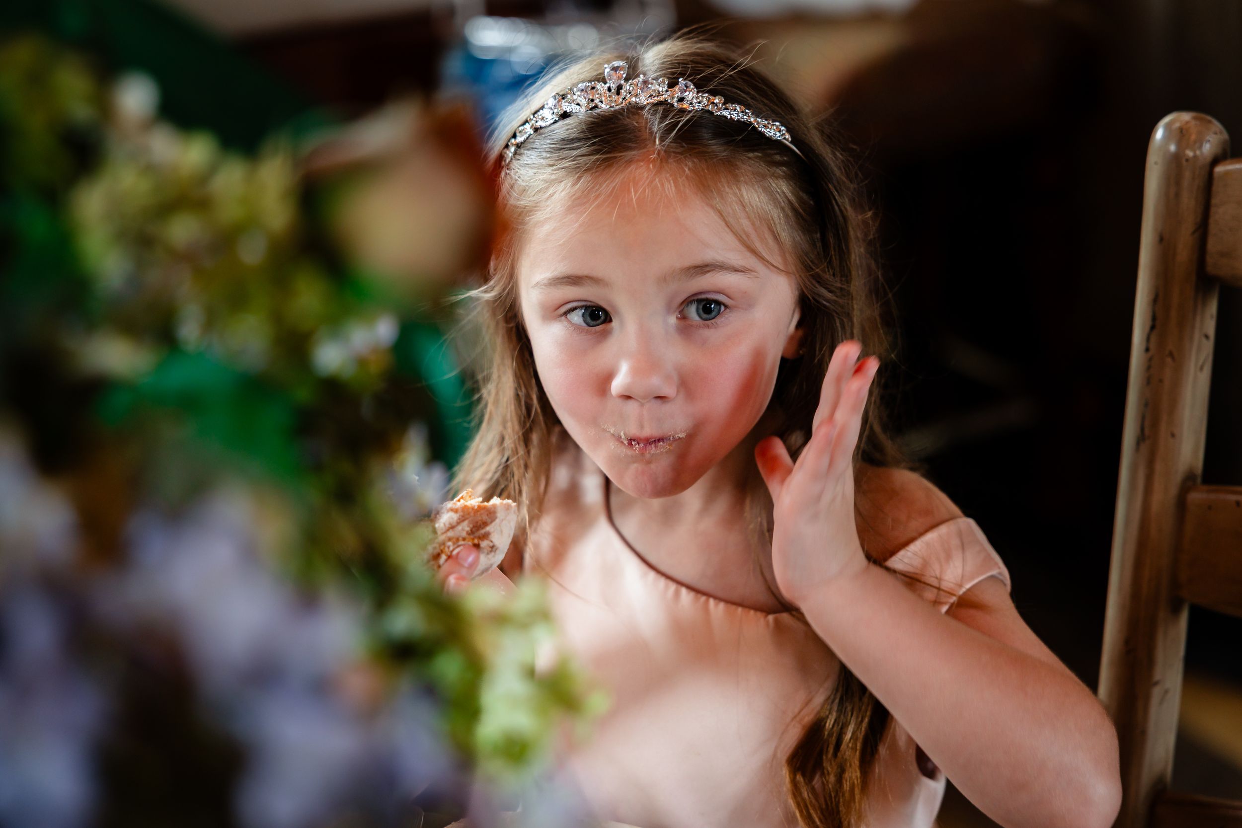 Adorable flower girl in tiara enjoying wedding reception treats at decorated table.