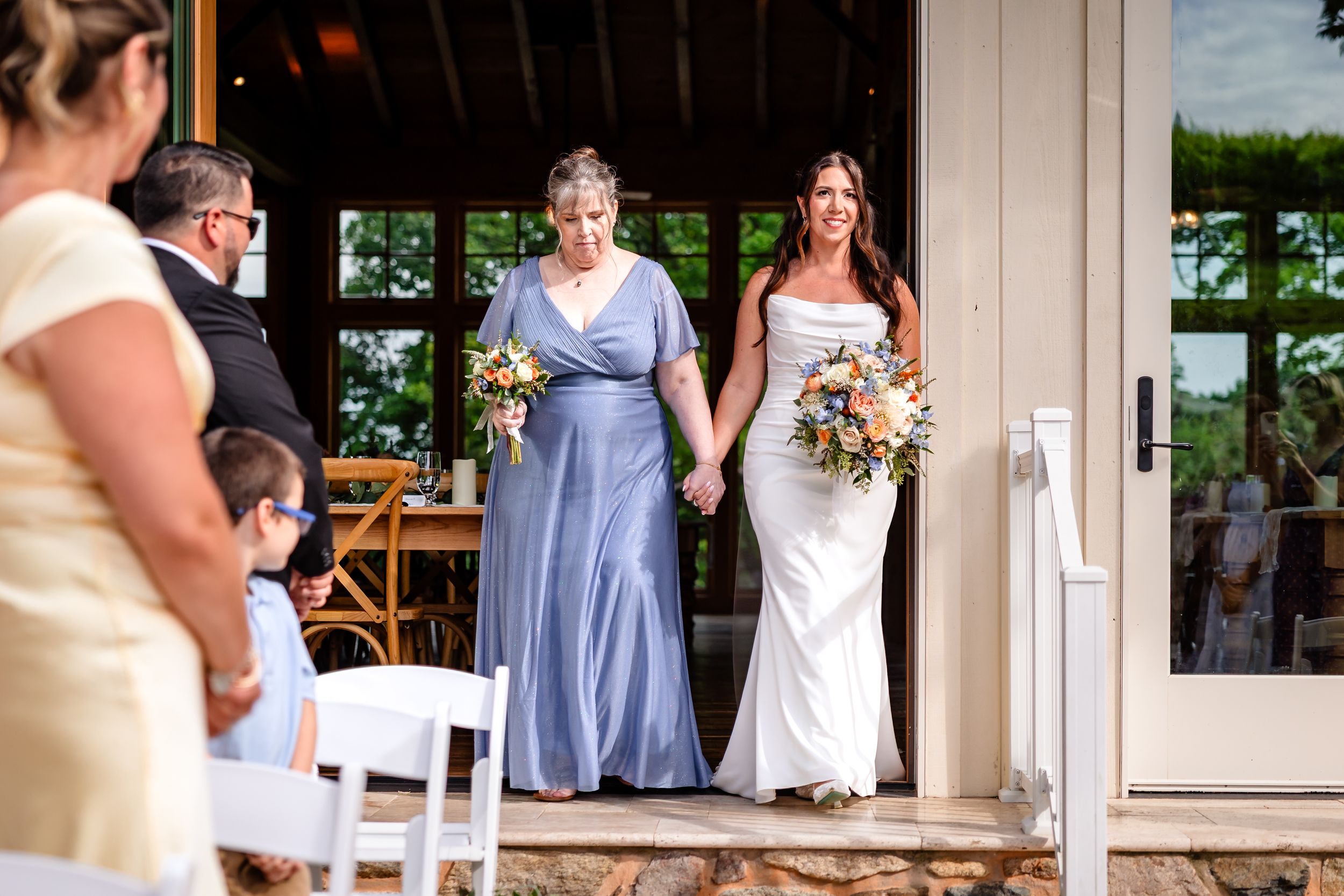 Bride in white gown walks with older woman in lavender dress during outdoor wedding ceremony.