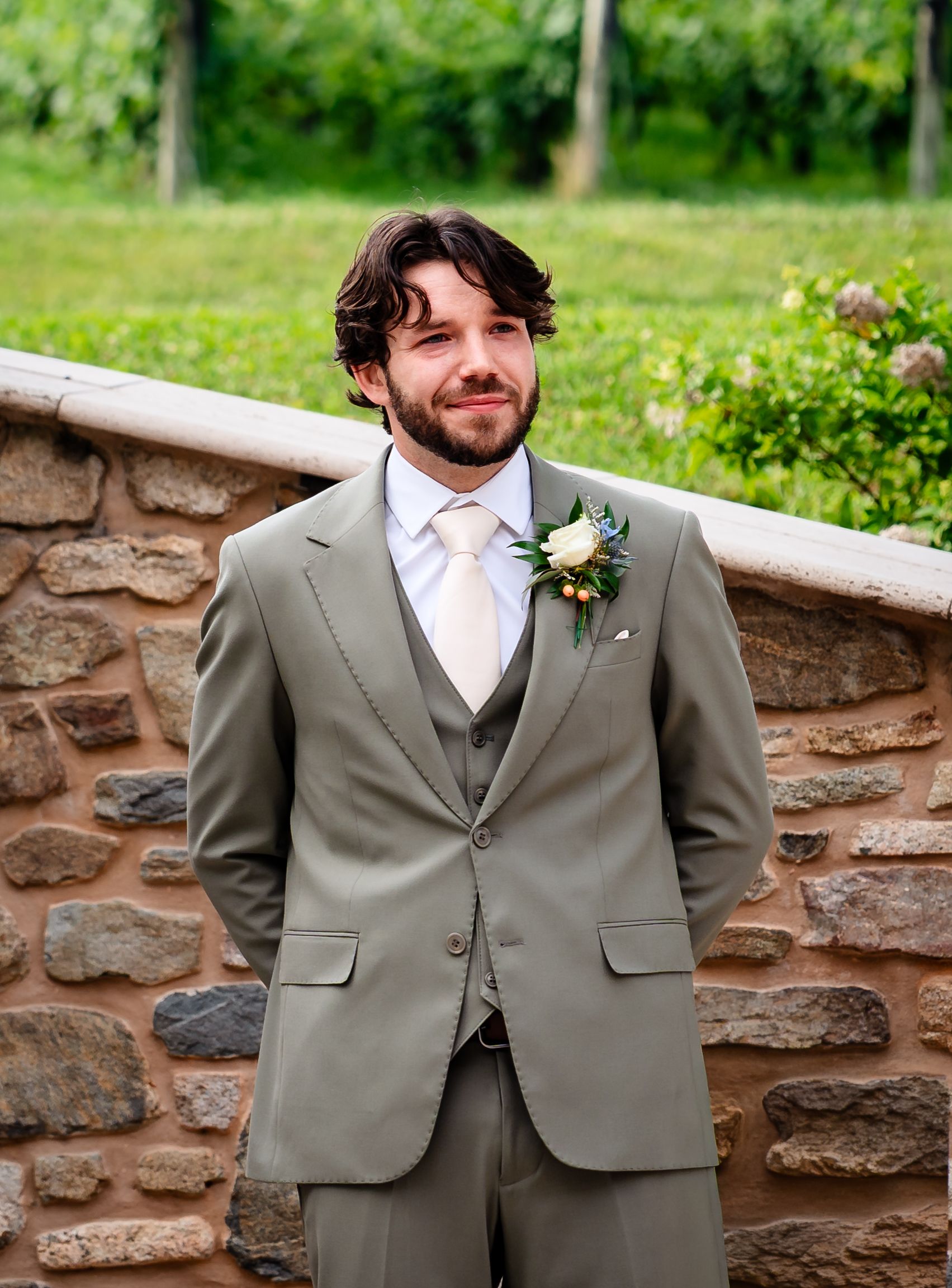 Groom in sage green three-piece suit with white boutonniere standing against rustic stone wall outdoors.