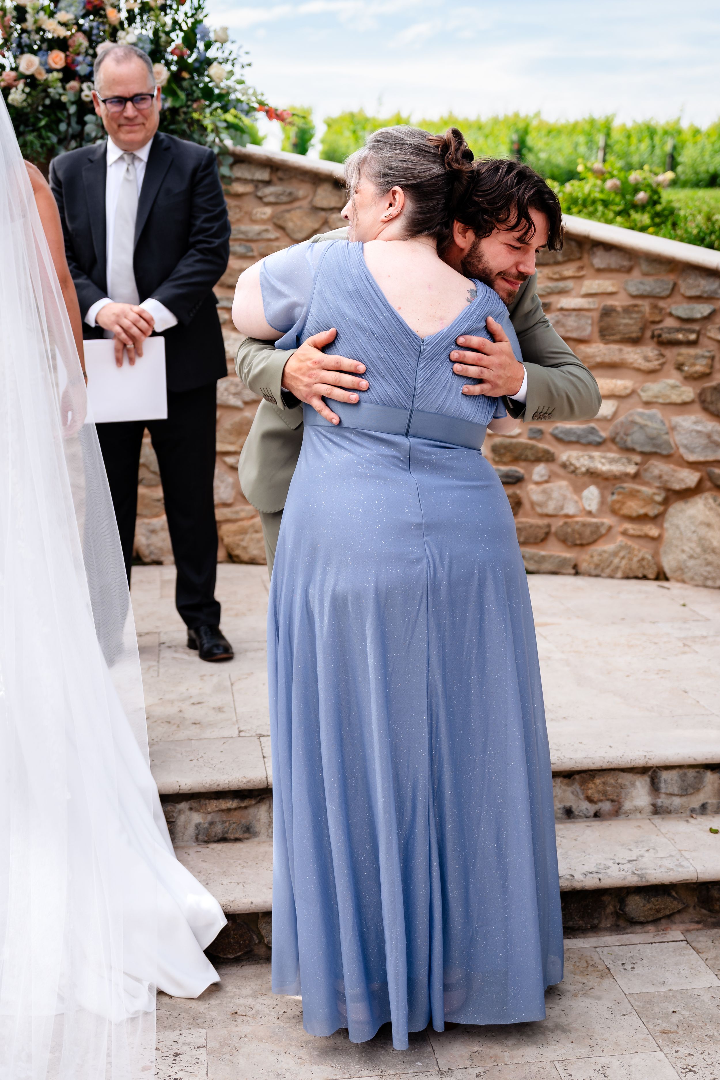 Groom embracing mother in blue gown at outdoor stone wedding ceremony venue.