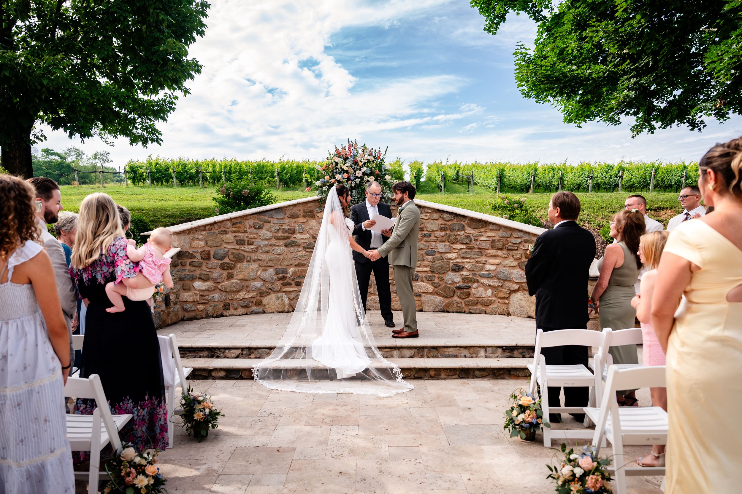 Bride and groom exchange vows at outdoor stone ceremony space with cornfield backdrop and guests seated.