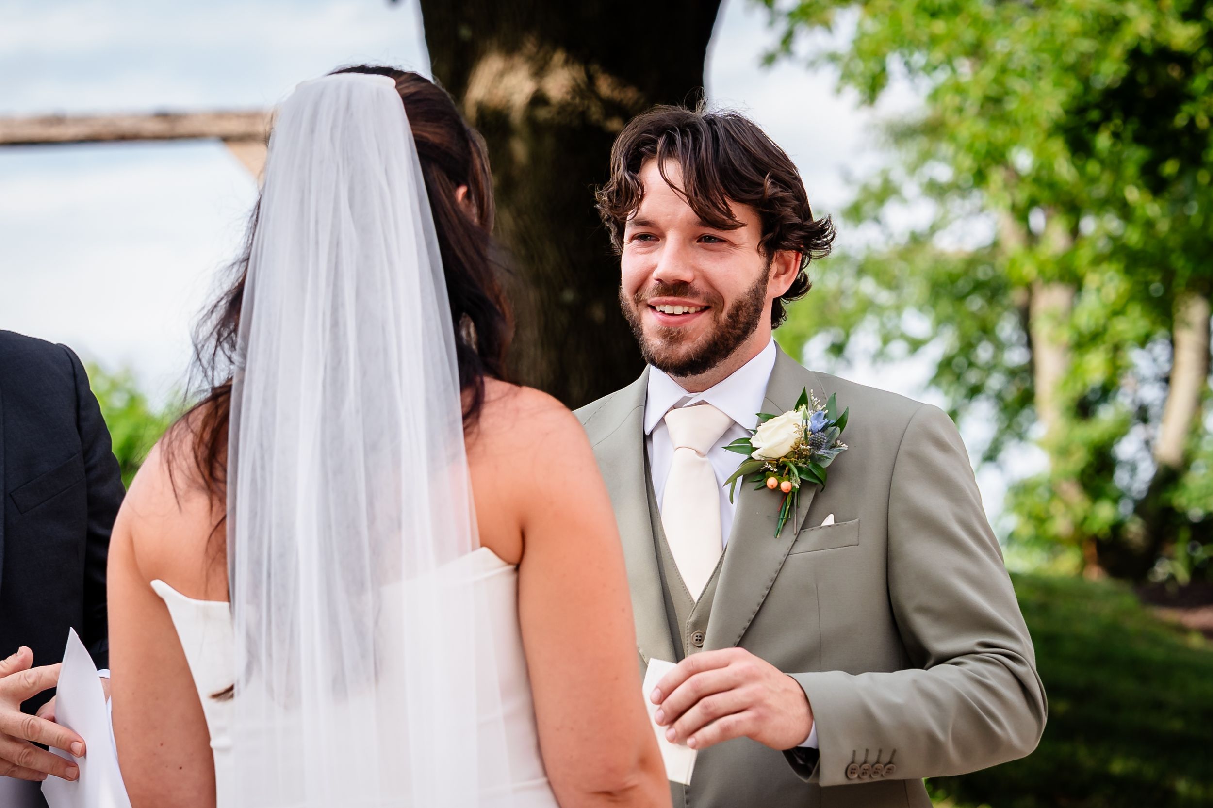Groom in sage suit smiles lovingly at veiled bride during outdoor wedding ceremony.