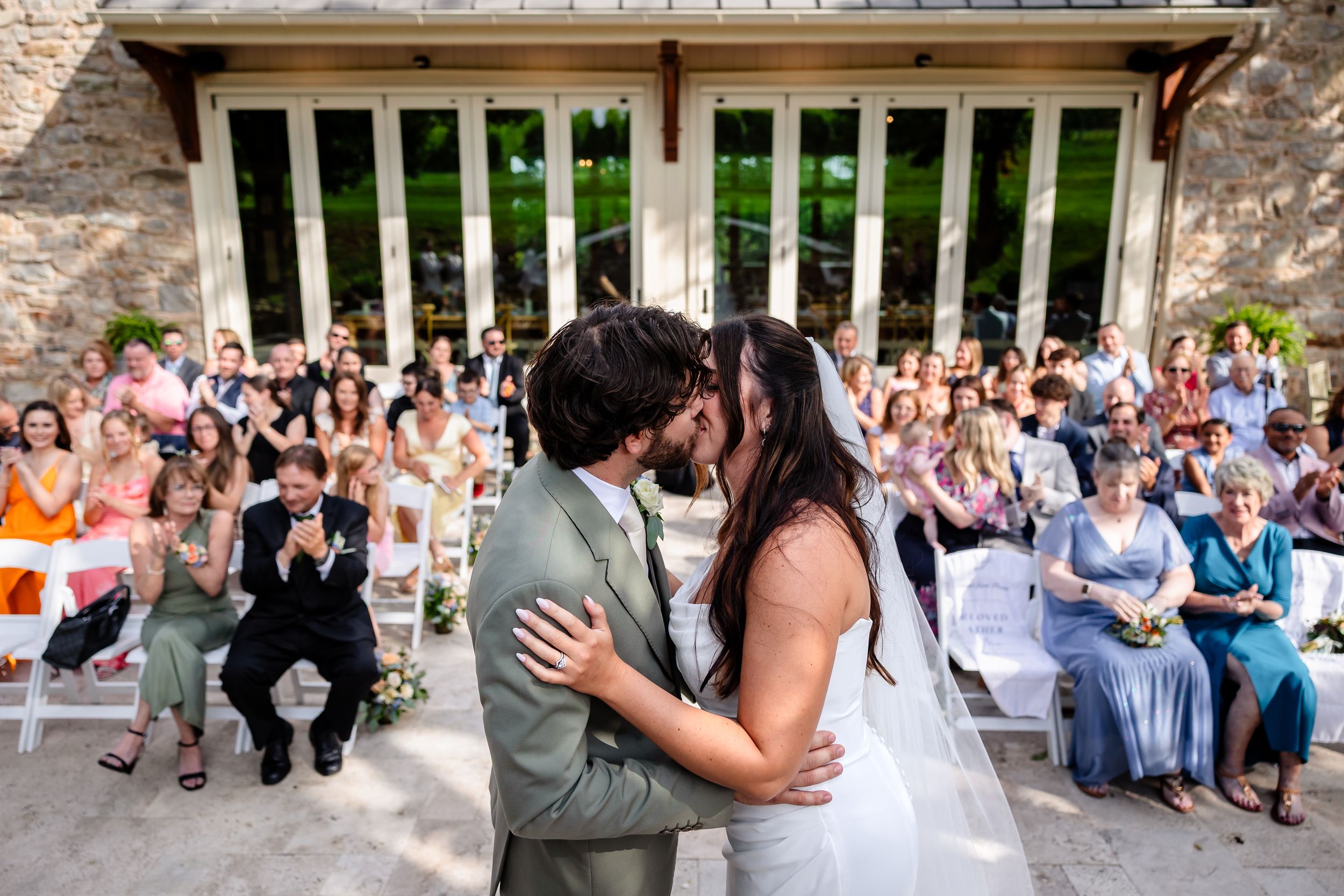 Bride and groom share first kiss at outdoor wedding ceremony with cheering guests behind.