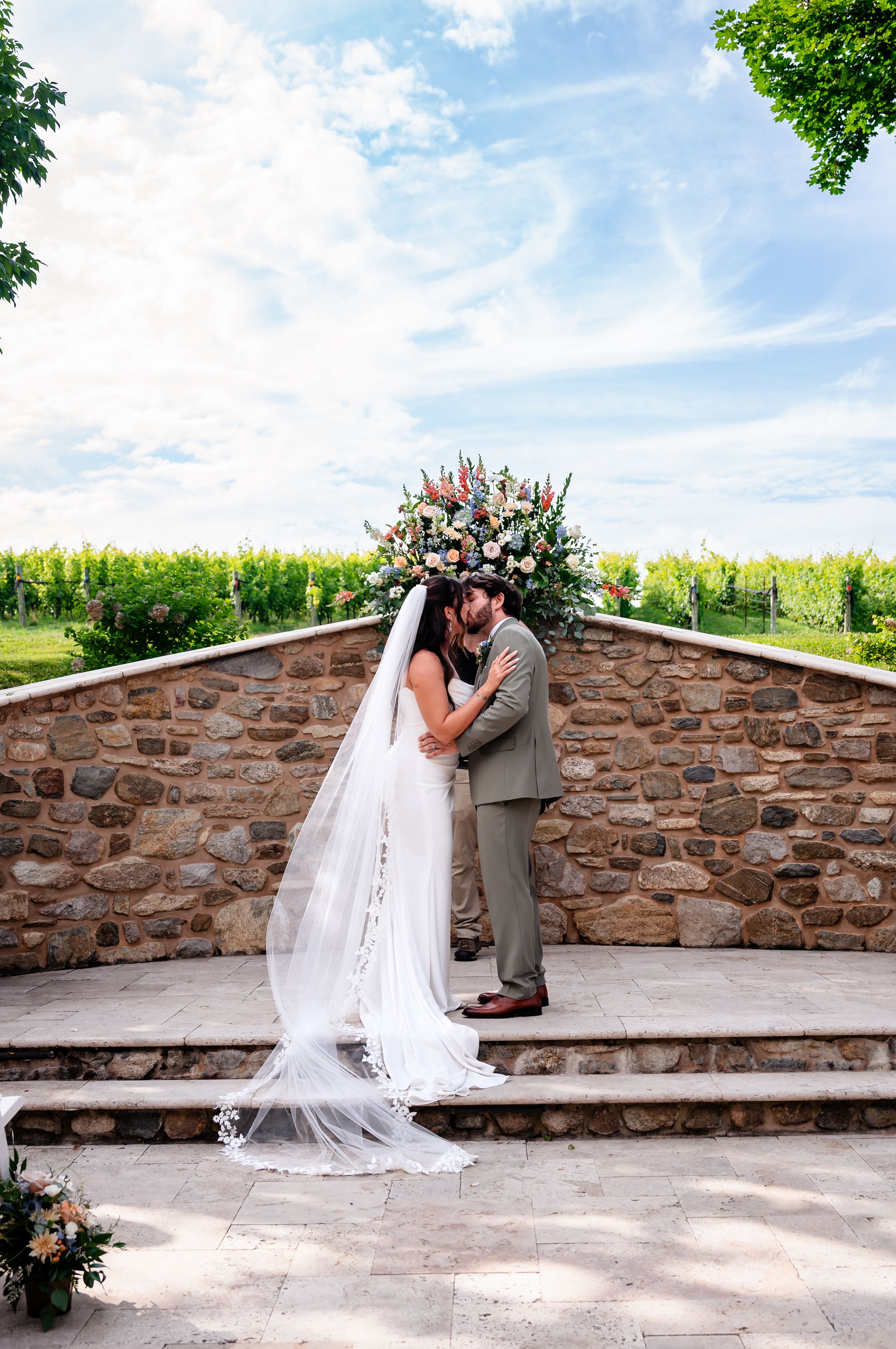 Bride and groom share first kiss at stone altar with floral arrangement under blue sky at vineyard.